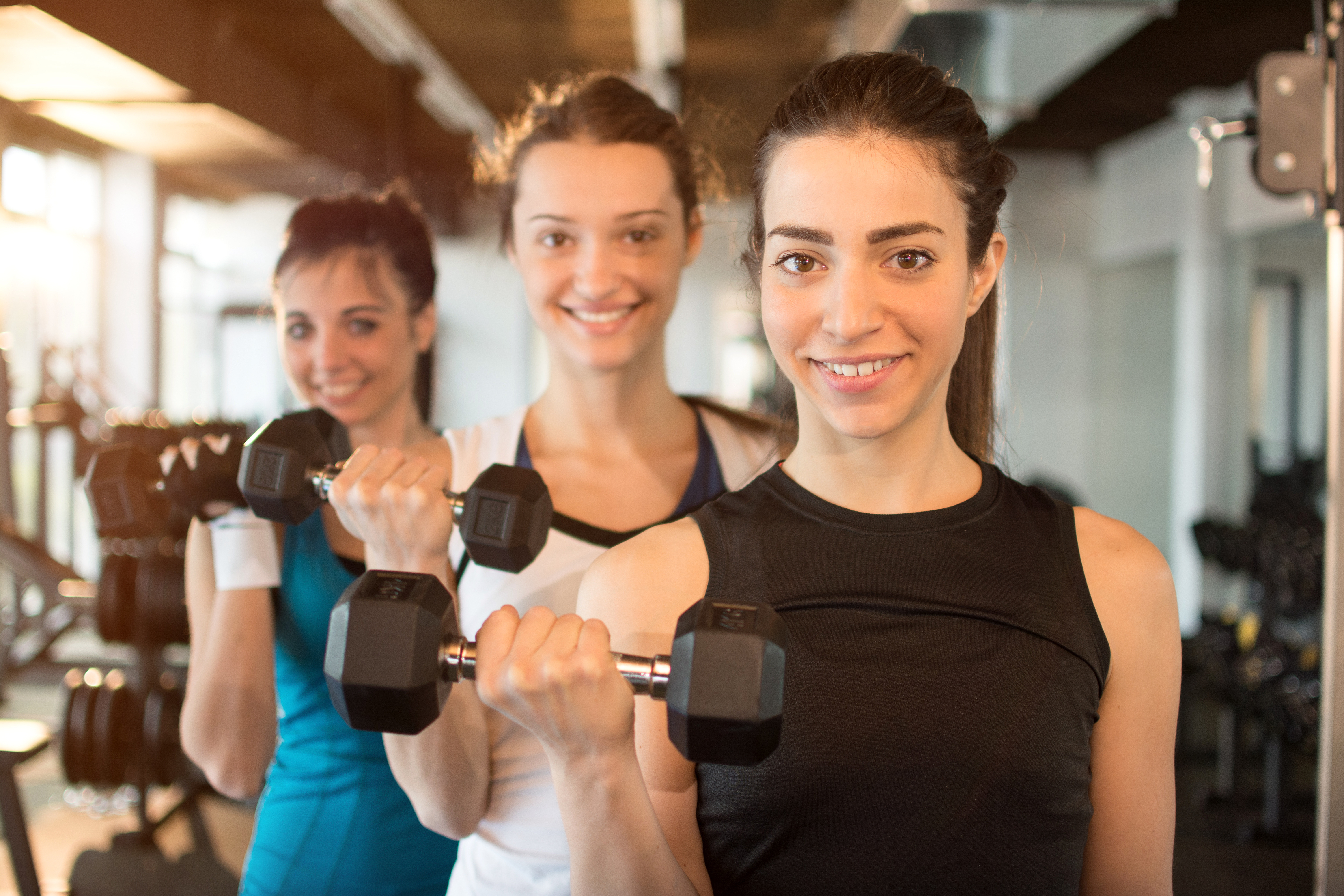 A group of people in a gym stand in a row performing bicep curls with dumbbells during a strength‑training session.