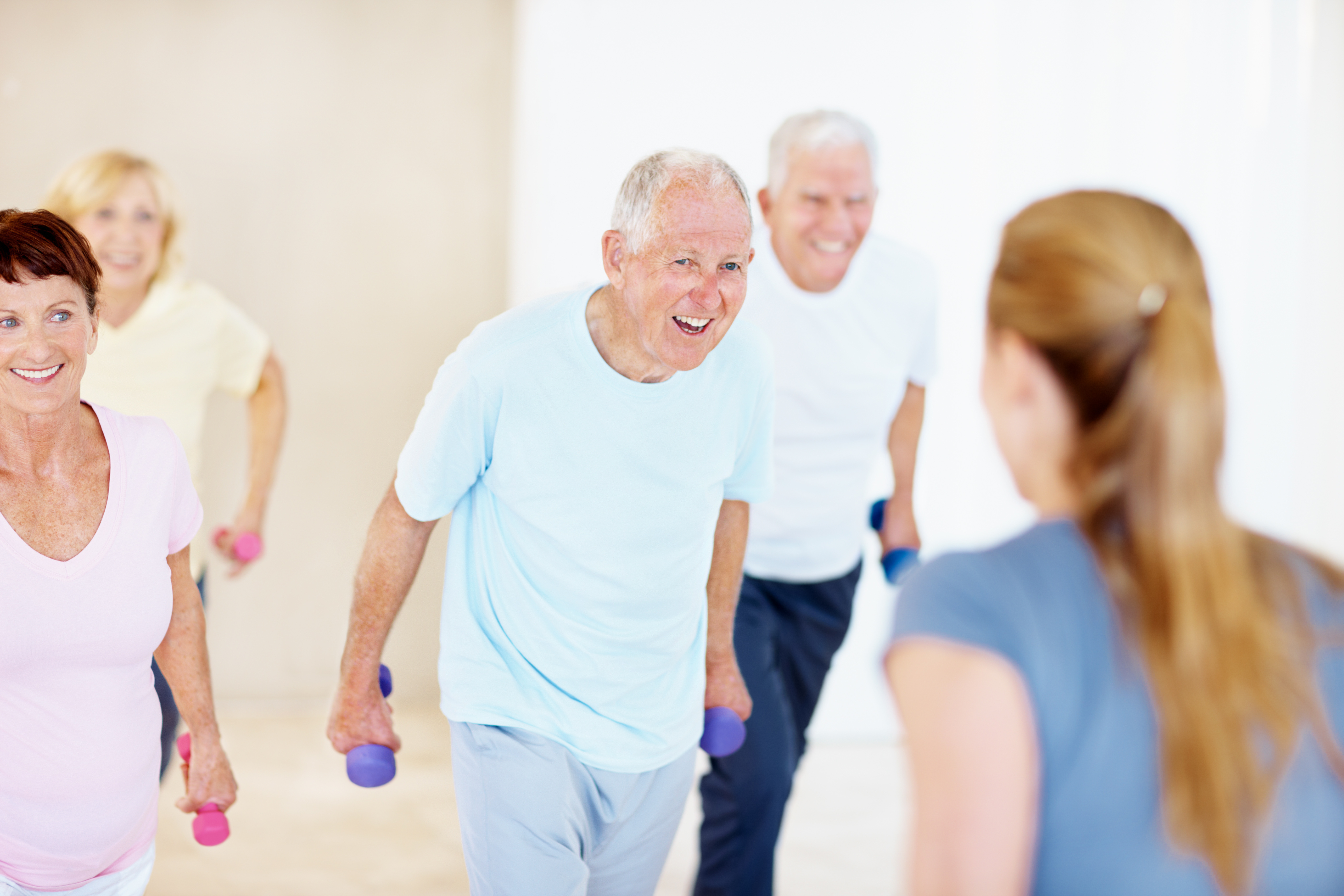 Smiling older adults holding small hand weights during an exercise class, with an instructor in the foreground.