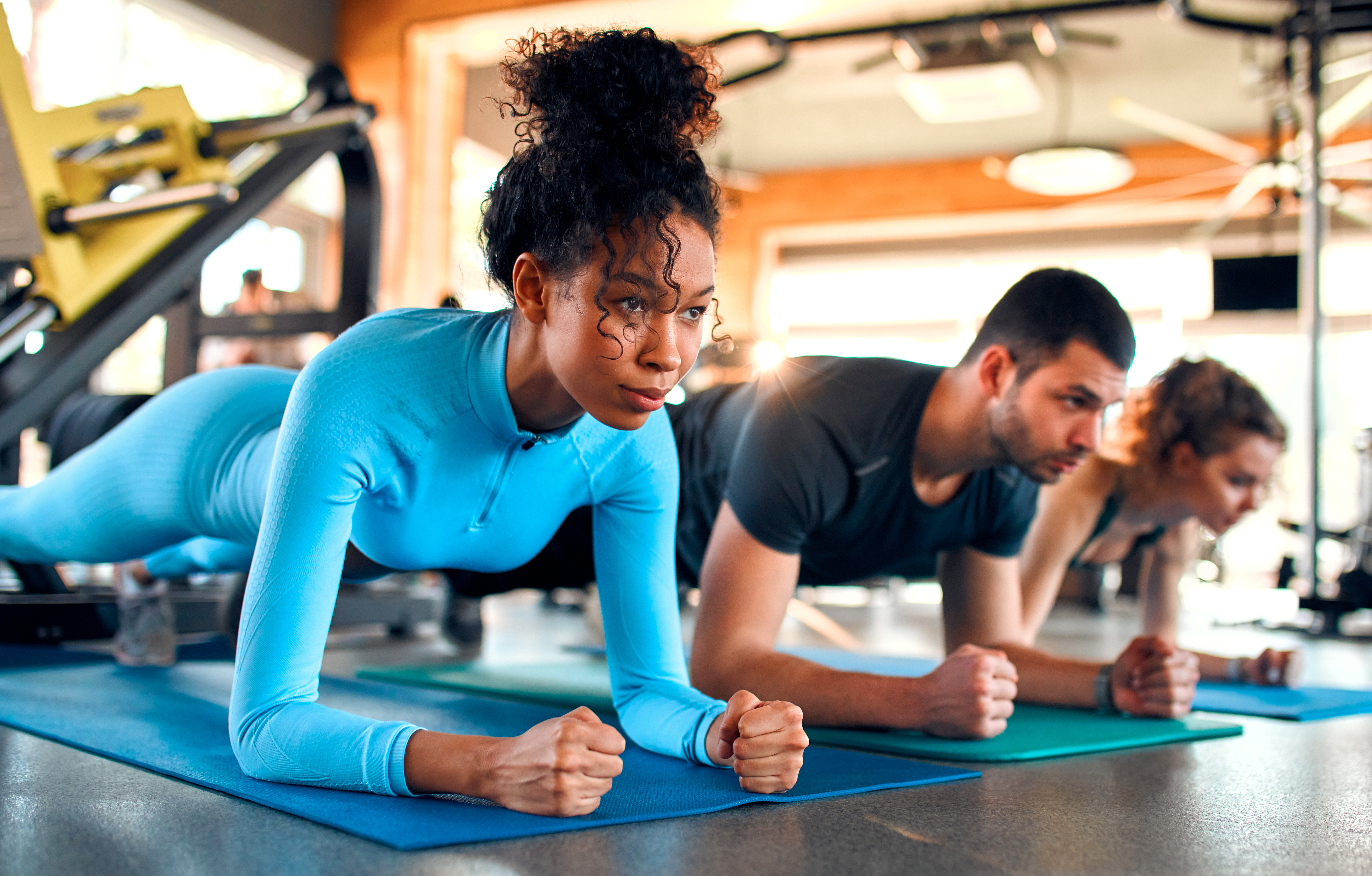 People exercising in a gym, holding a plank position on mats with fitness equipment in the background.