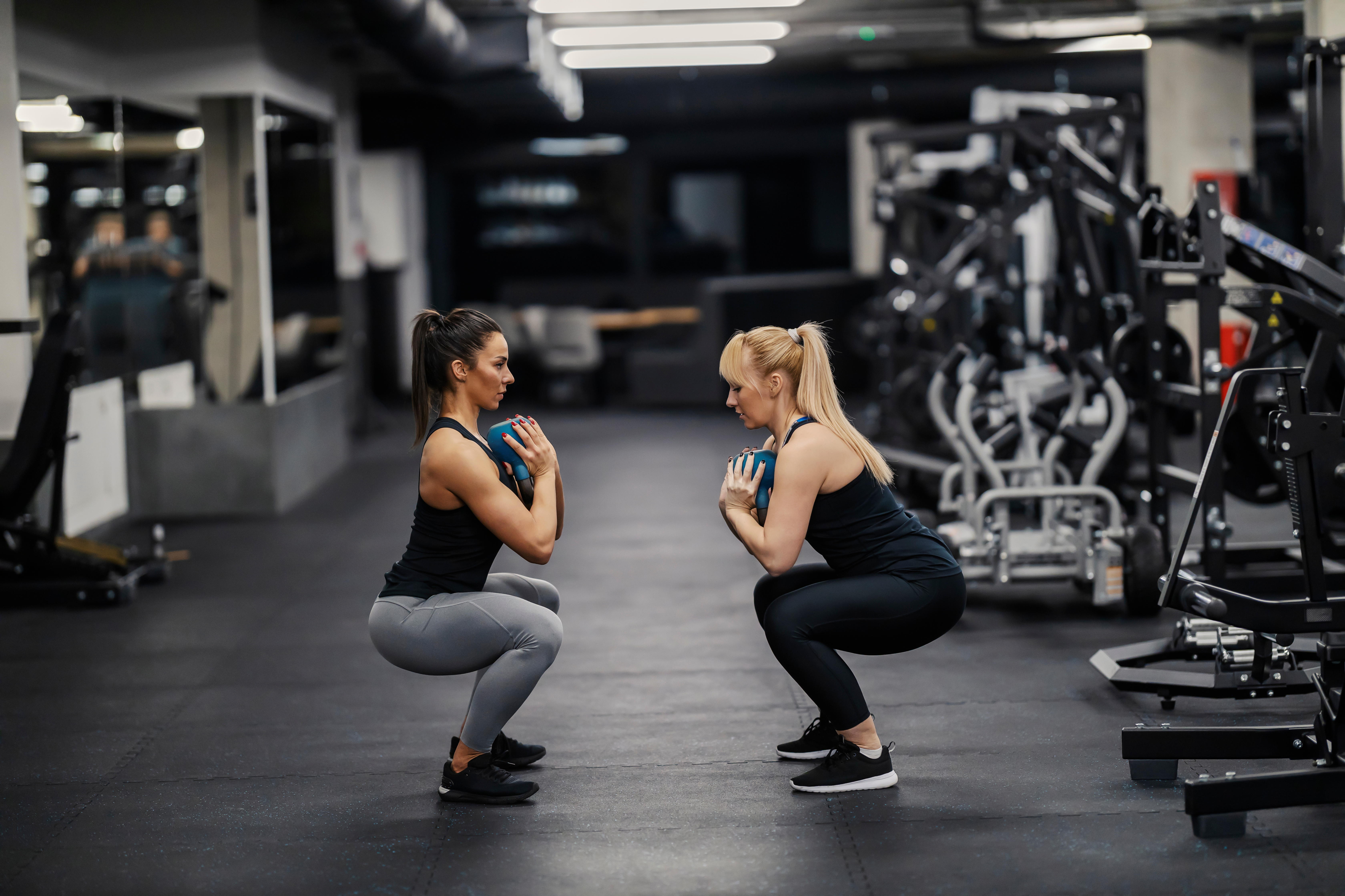 Two people performing weighted squats in a gym, each holding a small weight plate at their chest.