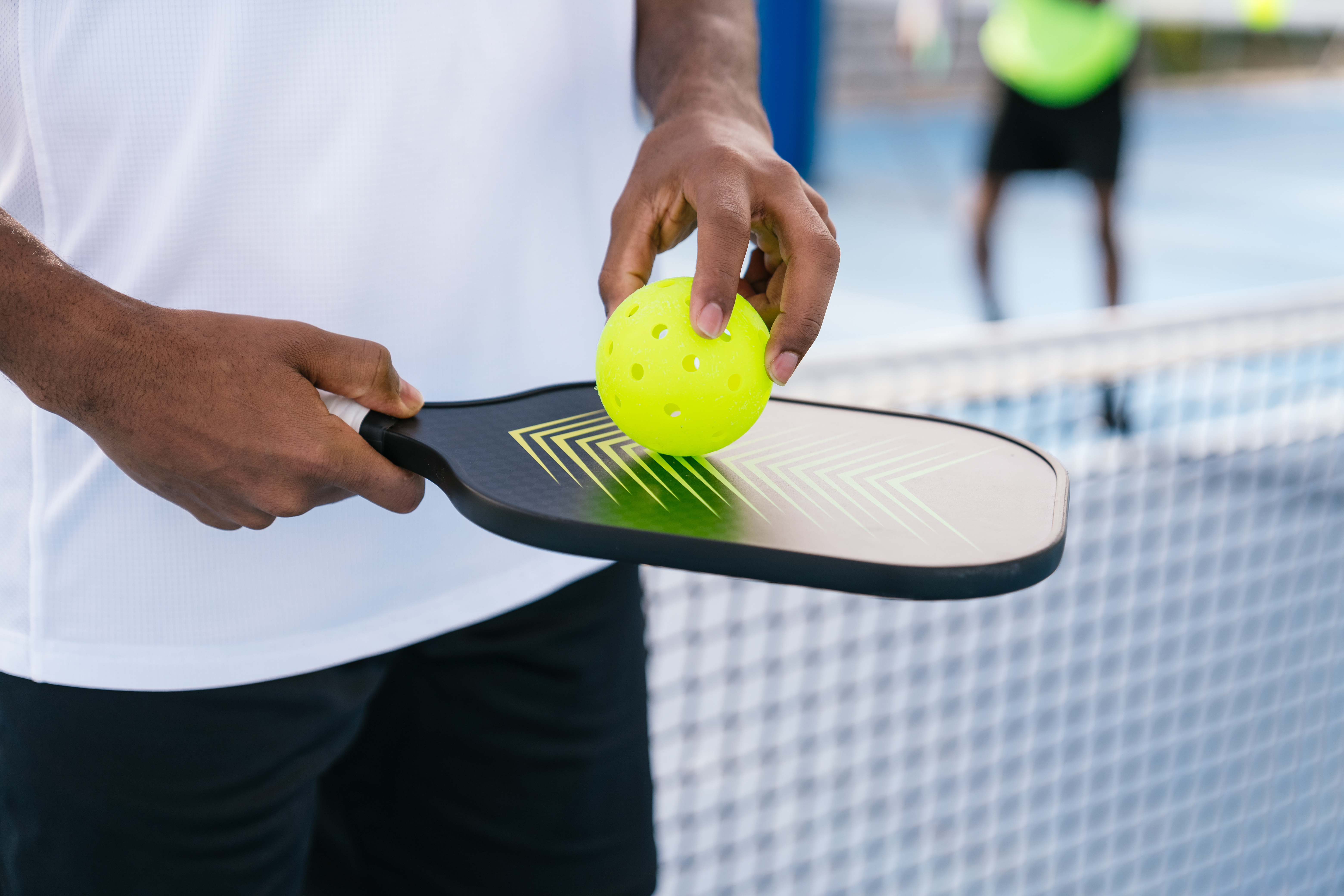 A person holds a yellow pickleball on a paddle at the edge of a court.