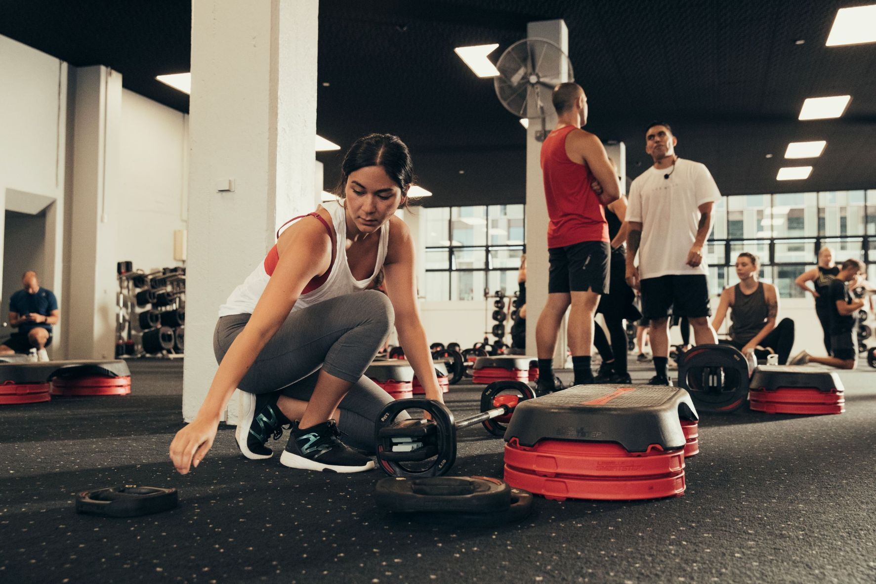 People in a gym getting ready for a group strength class, setting up barbells and step platforms on the floor.
