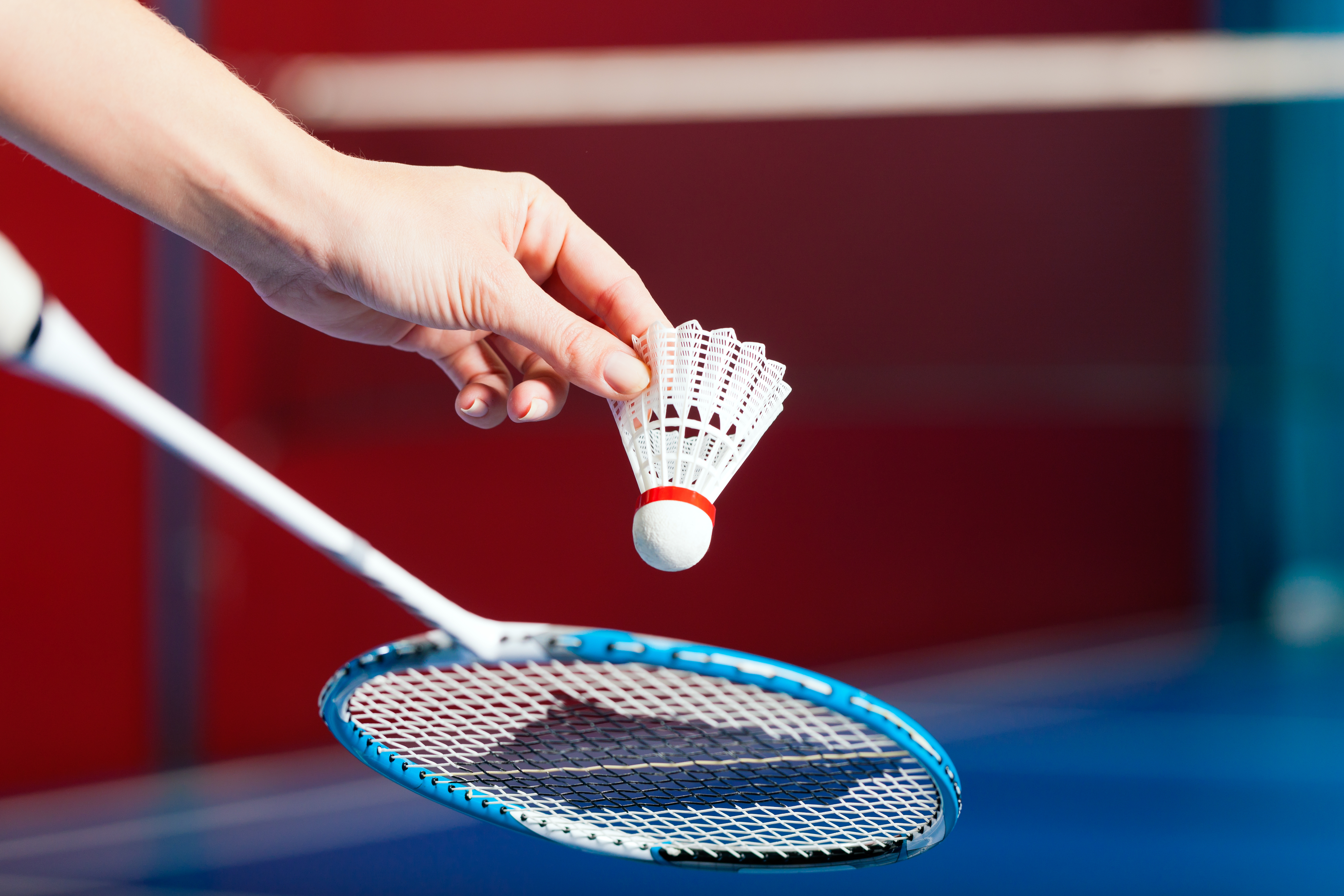 A hand holds a shuttlecock above a badminton racket on a court.