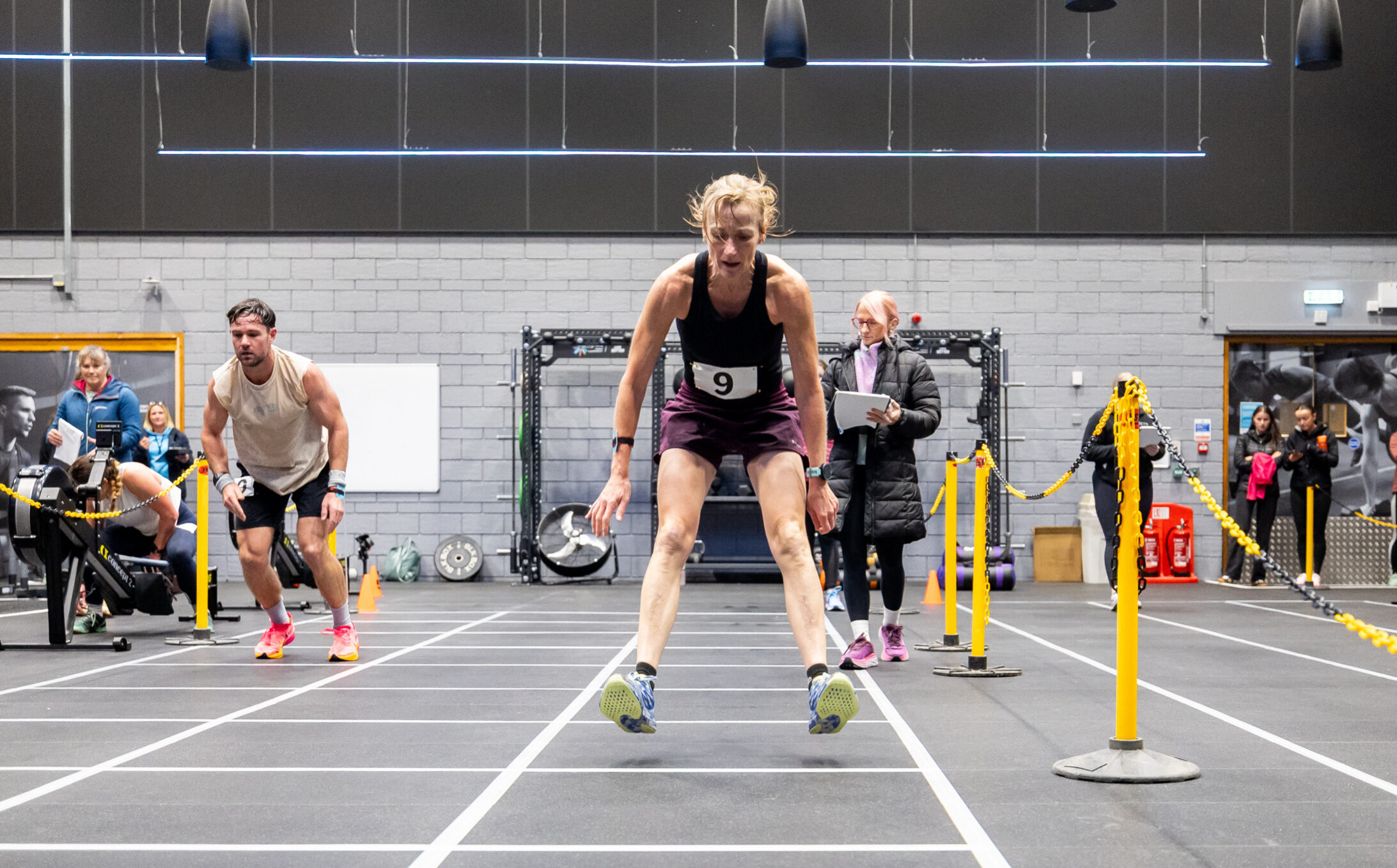 A person competing in an indoor fitness race, jumping forward between marked lanes while others and event officials watch in the background.