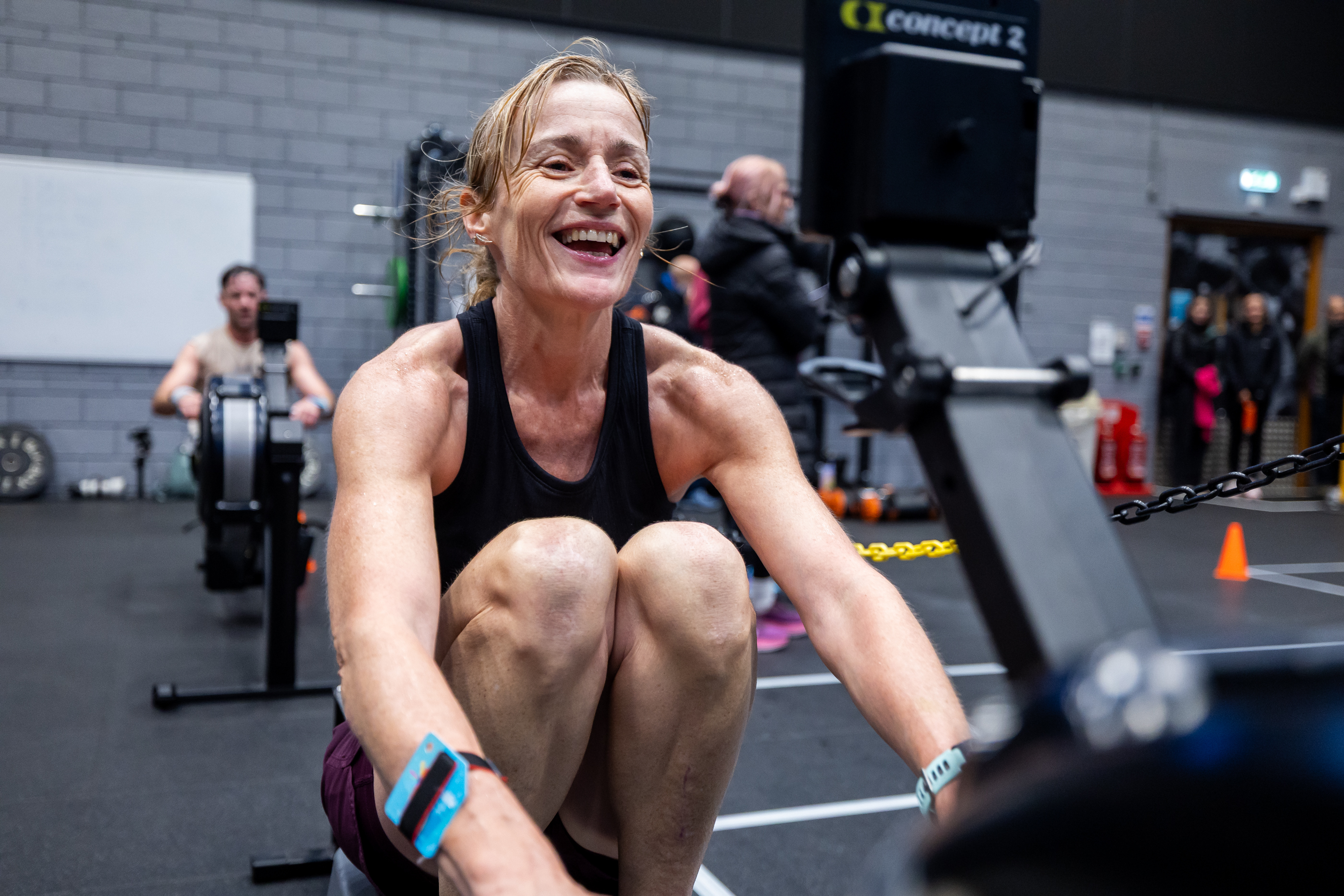 Person using a rowing machine during an indoor fitness event, leaning forward in the rowing motion with knees bent and arms extended. The scene takes place in a gym hall with other participants and spectators in the background, along with fitness equipment and marked exercise areas.