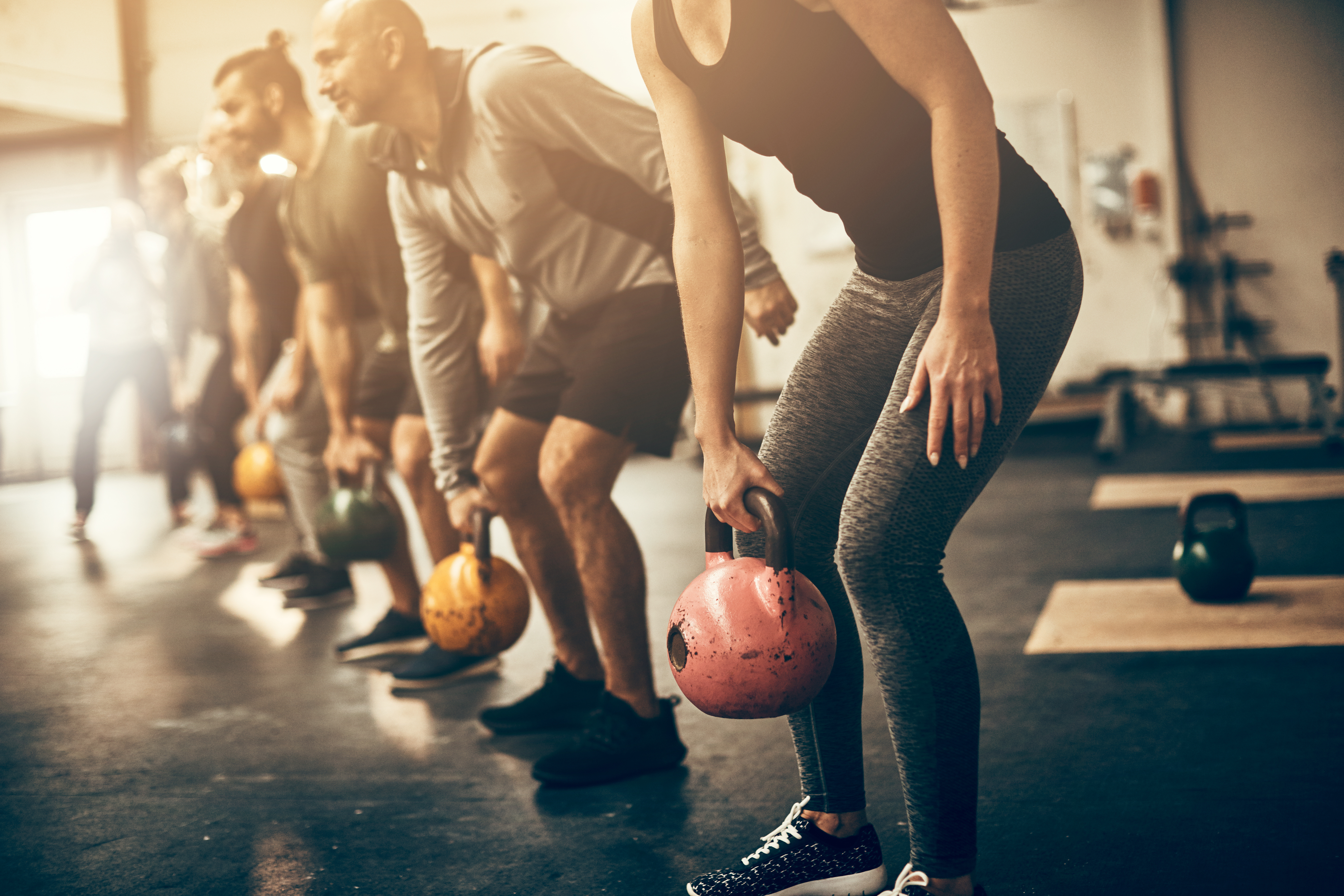Group fitness class performing kettlebell exercises in a gym, with participants lined up and bending forward while holding colorful kettlebells on a dark floor.