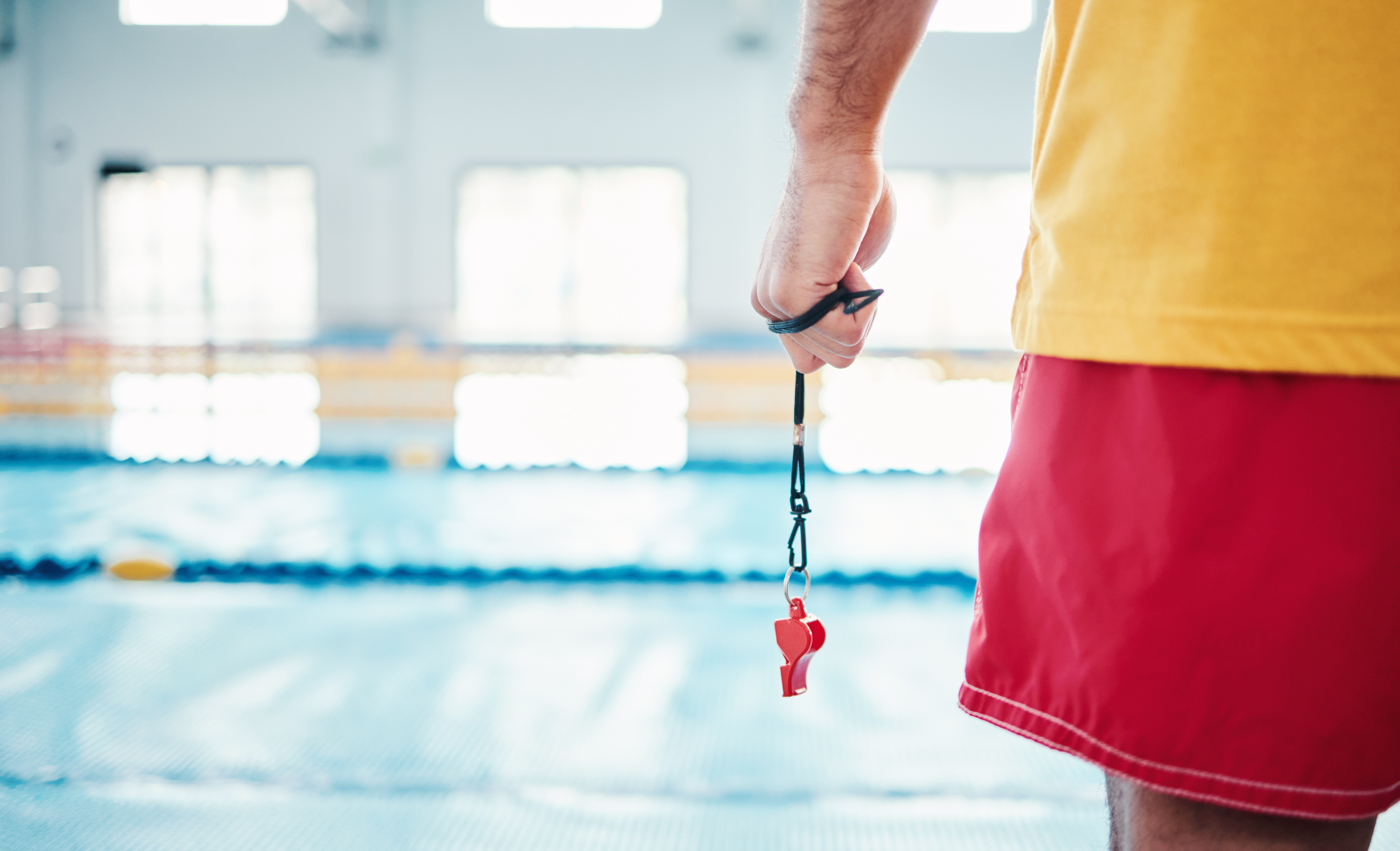 A lifeguard standing beside a swimming pool holding a whistle on a lanyard.