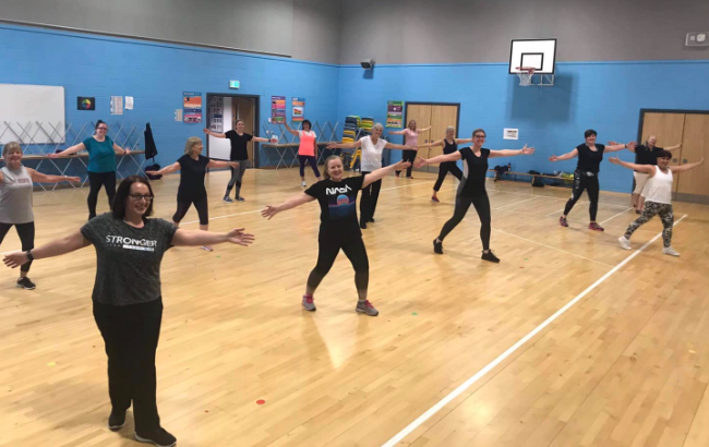 A group fitness class in a sports hall, with participants standing in a wide stance and stretching their arms out to the sides.