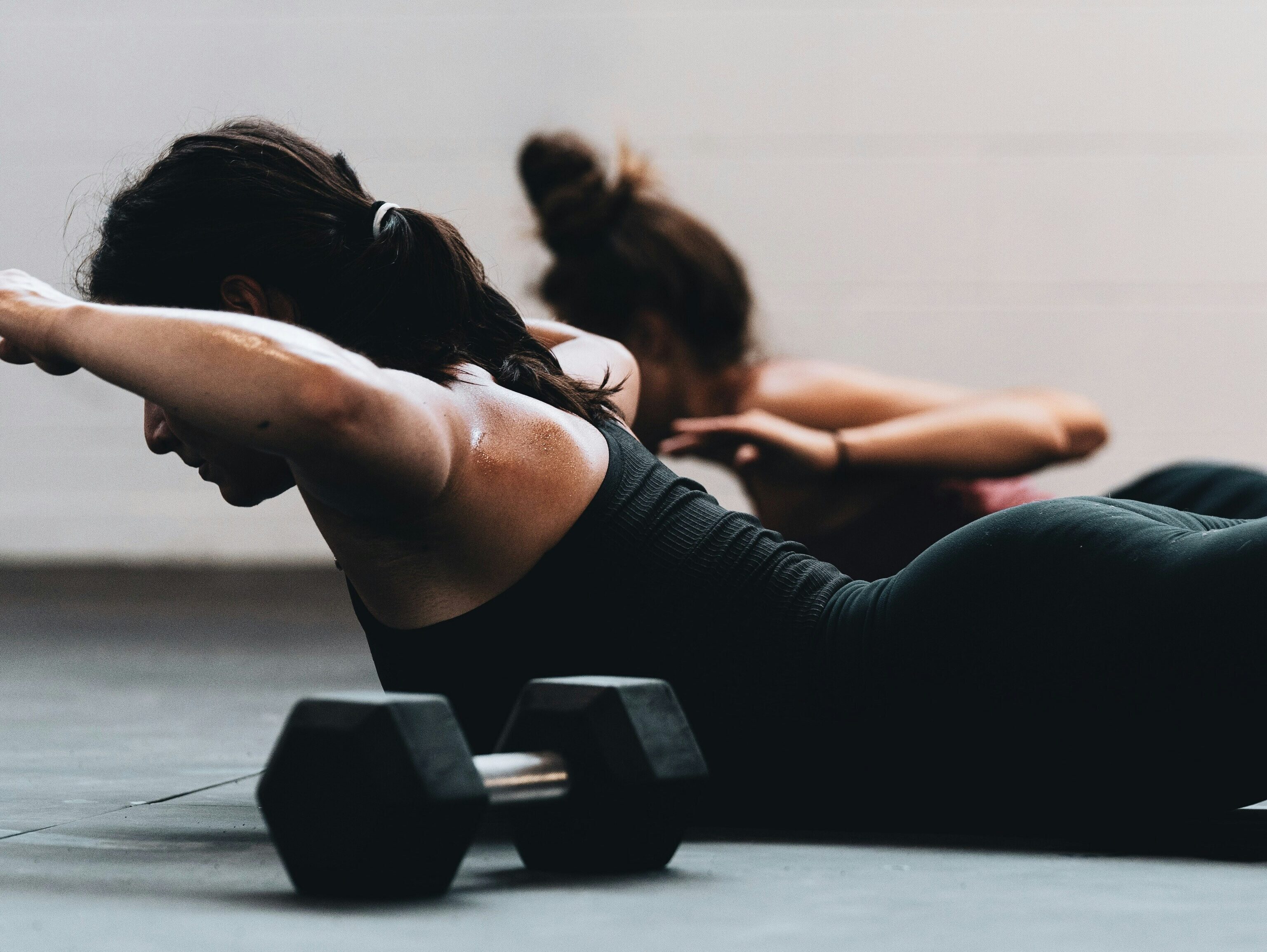 Two people lying on the floor in a gym, lifting their arms and upper bodies in a back‑strengthening exercise beside a set of dumbbells.