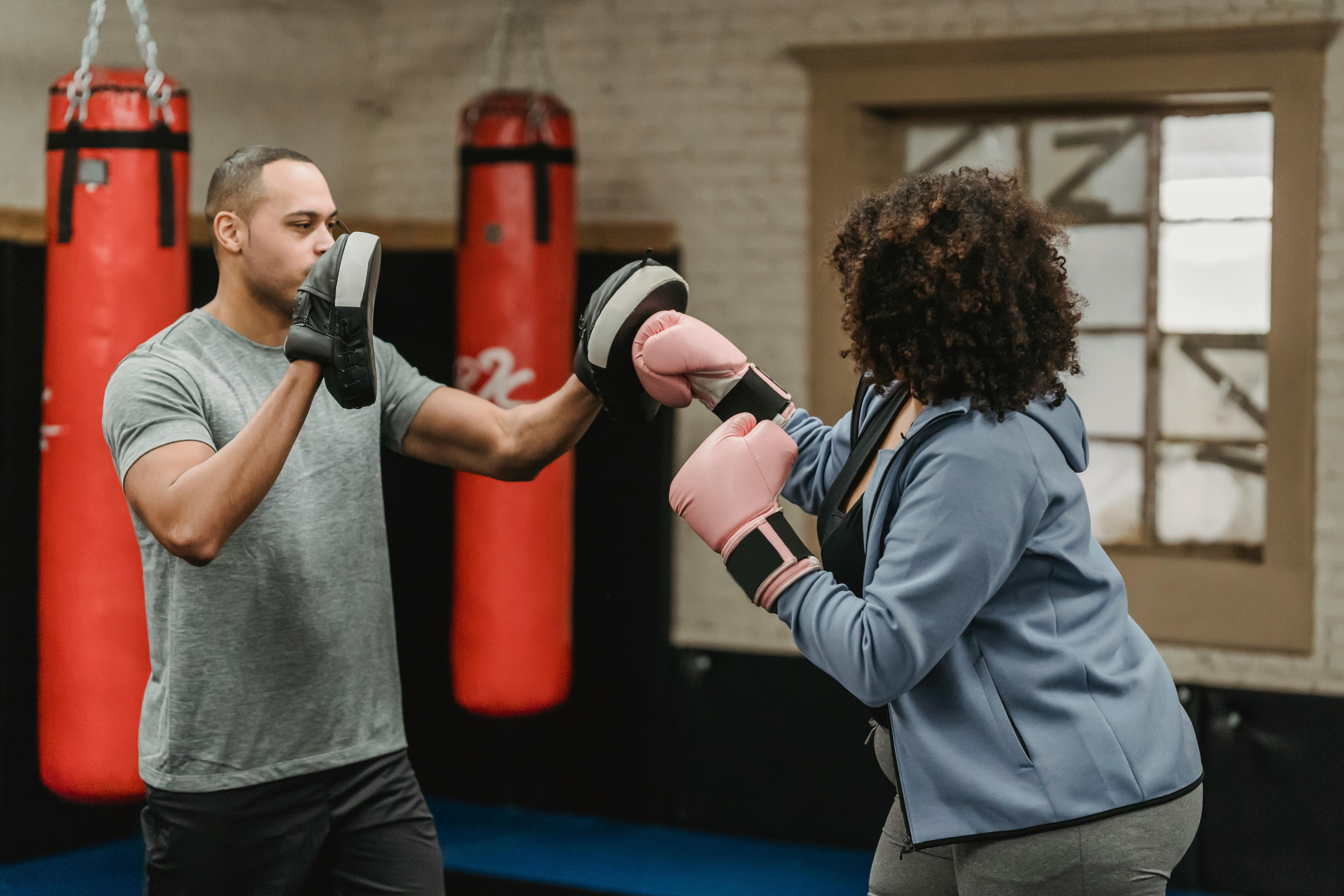 A boxing trainer holding focus pads while a person wearing pink gloves practises punches in a gym with heavy bags in the background.