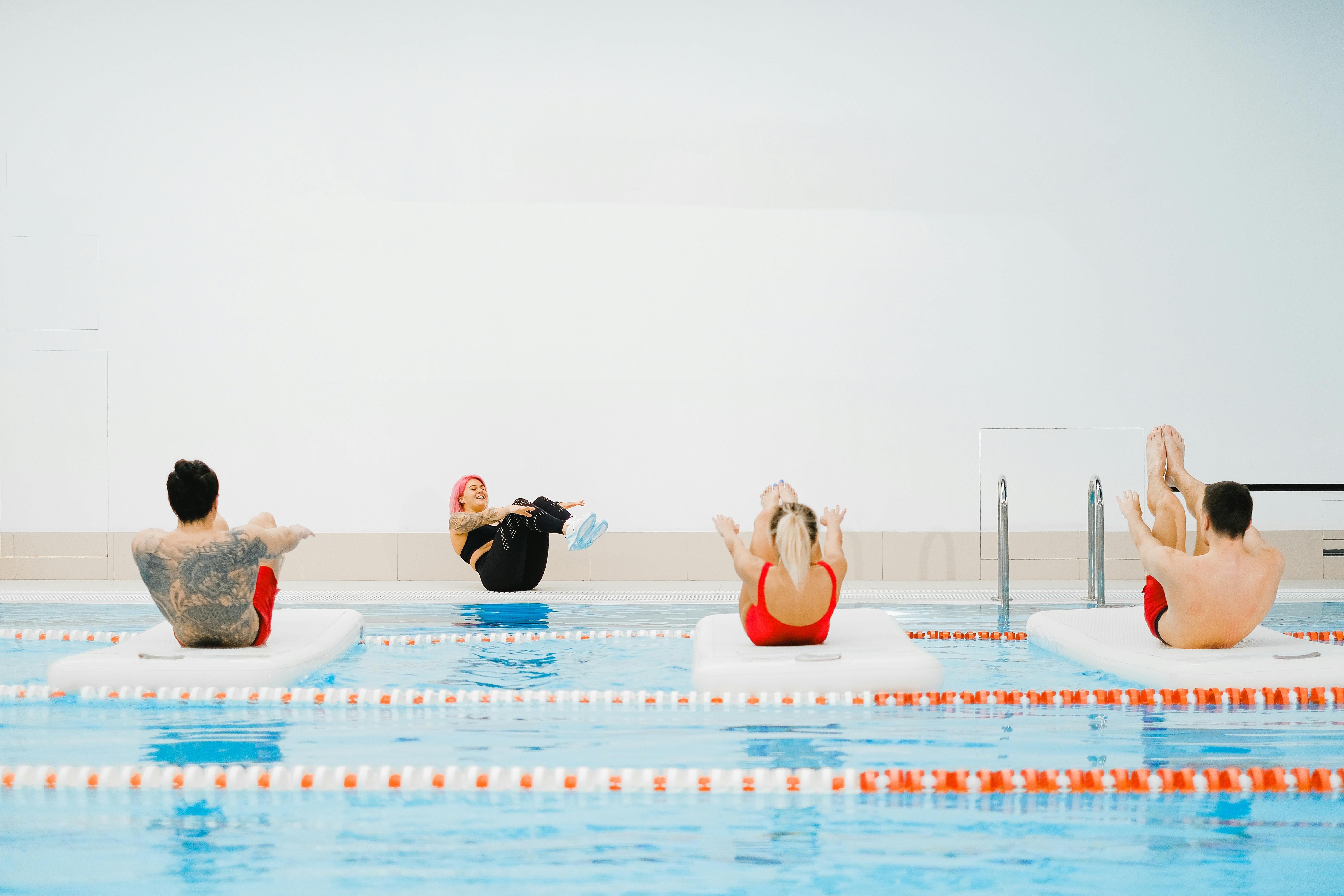 People taking part in a pool-based fitness class, balancing on floating mats while following an instructor at the pool edge.