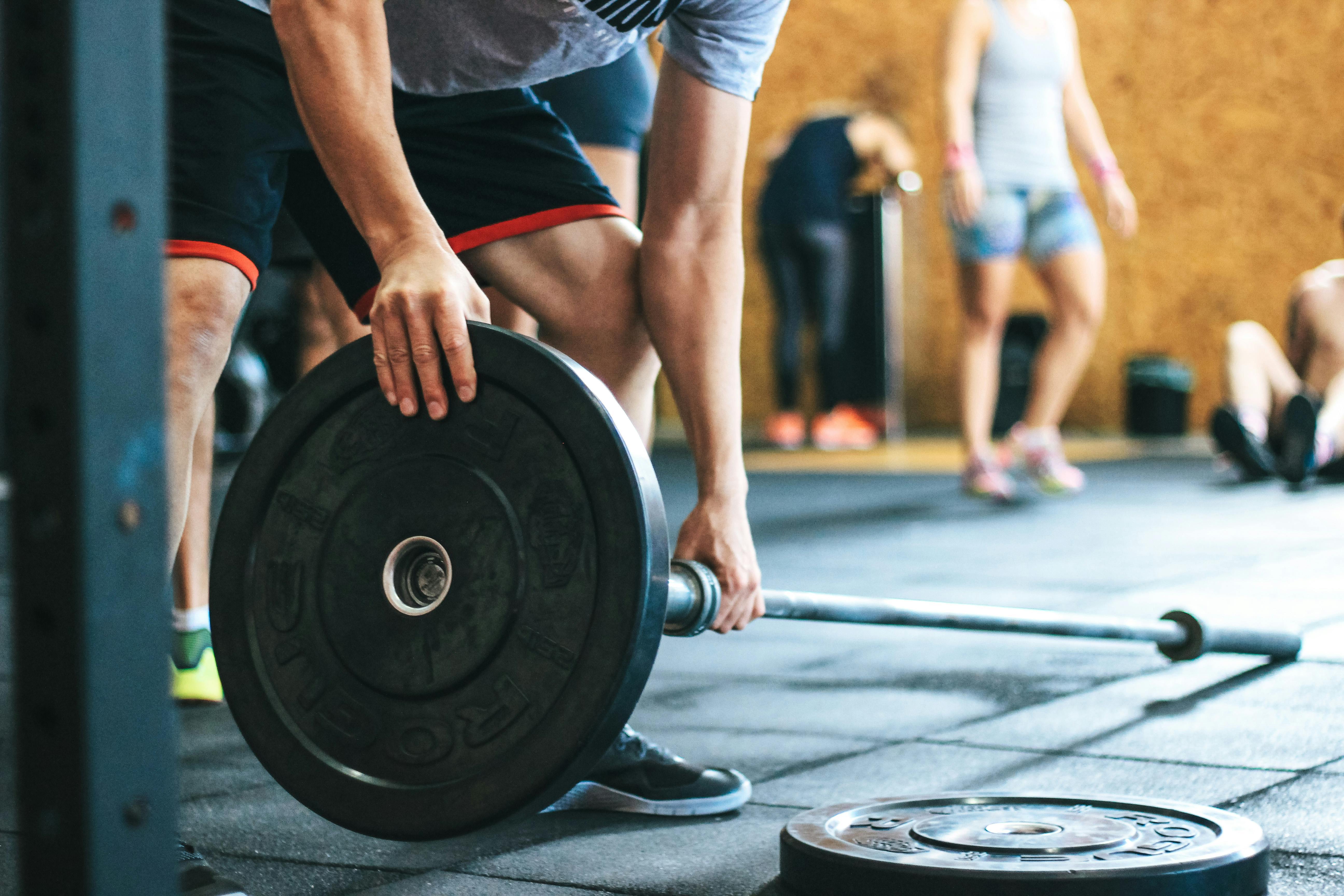 A person preparing to lift a barbell by adding weight plates in a gym, with others training in the background.