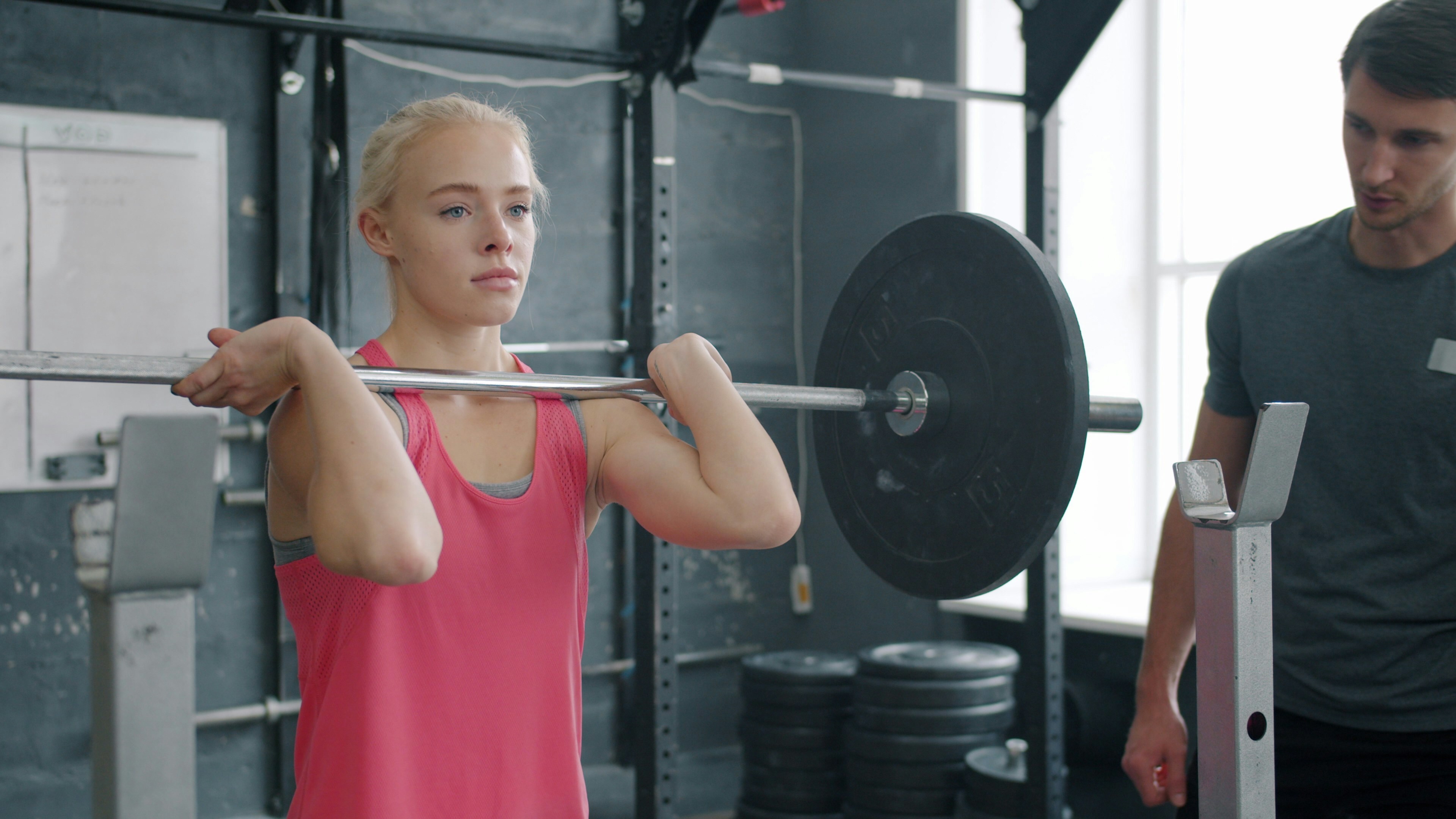 A person lifting a barbell in a gym while another person stands nearby providing support.