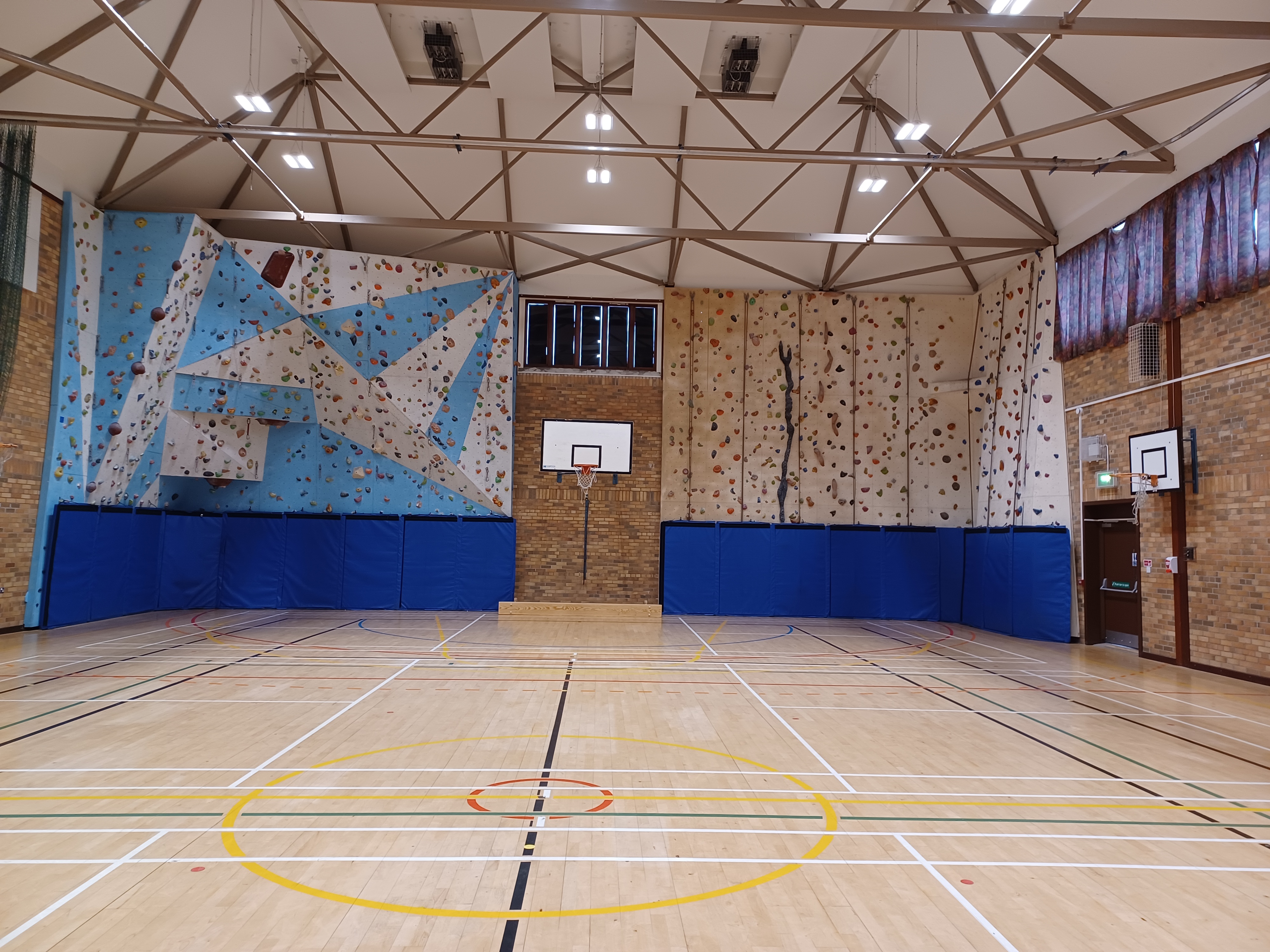 Spacious indoor games hall with a polished wooden floor marked for multiple sports, featuring two colorful climbing walls and a basketball hoop mounted on a brick wall.