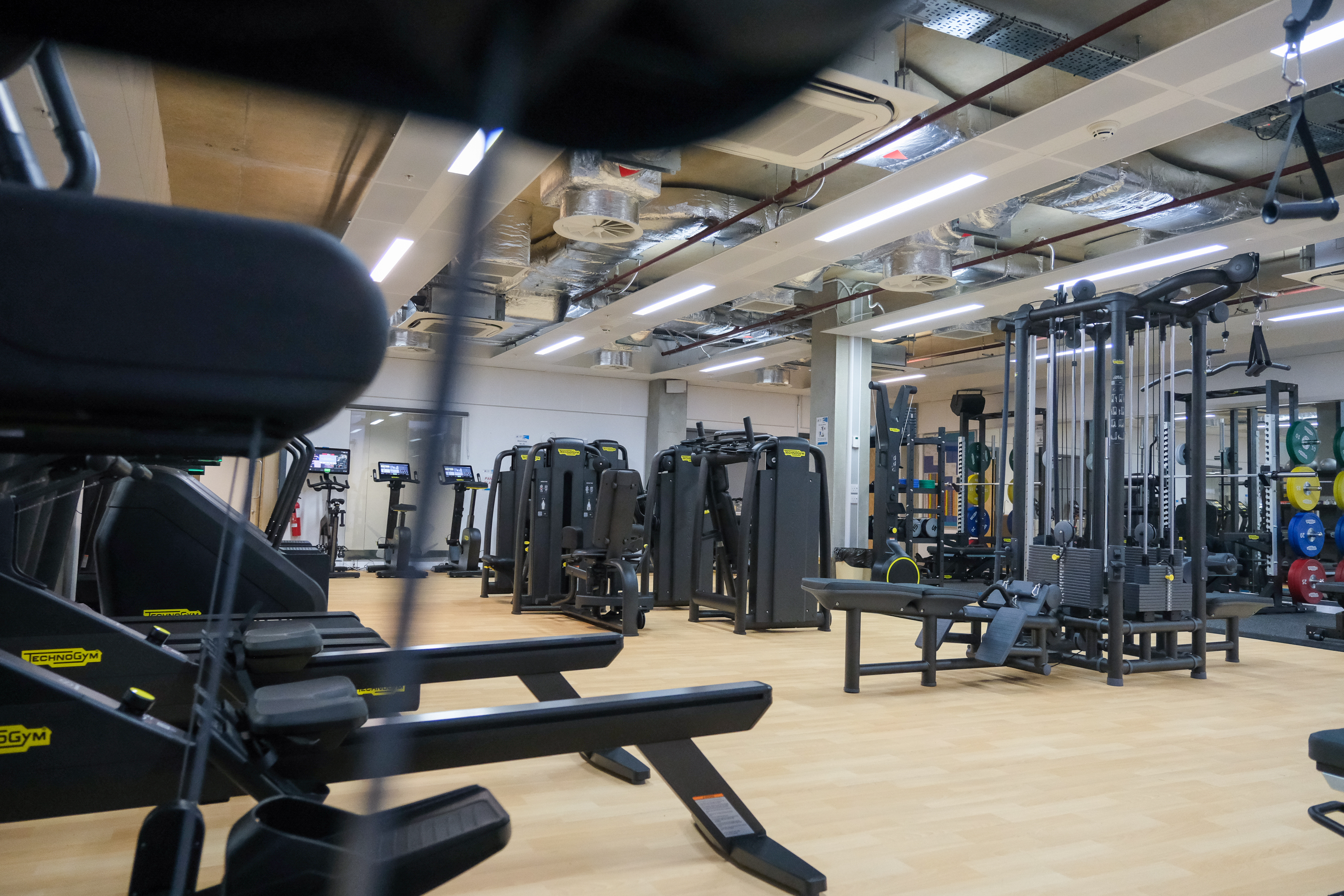 Spacious modern gym with light wood flooring and rows of black strength‑training machines, cable stations, and benches. Cardio equipment is visible in the background, and weight plates are neatly stored on racks. Overhead, exposed ducting and bright strip lighting run across the ceiling, creating an industrial, open feel.