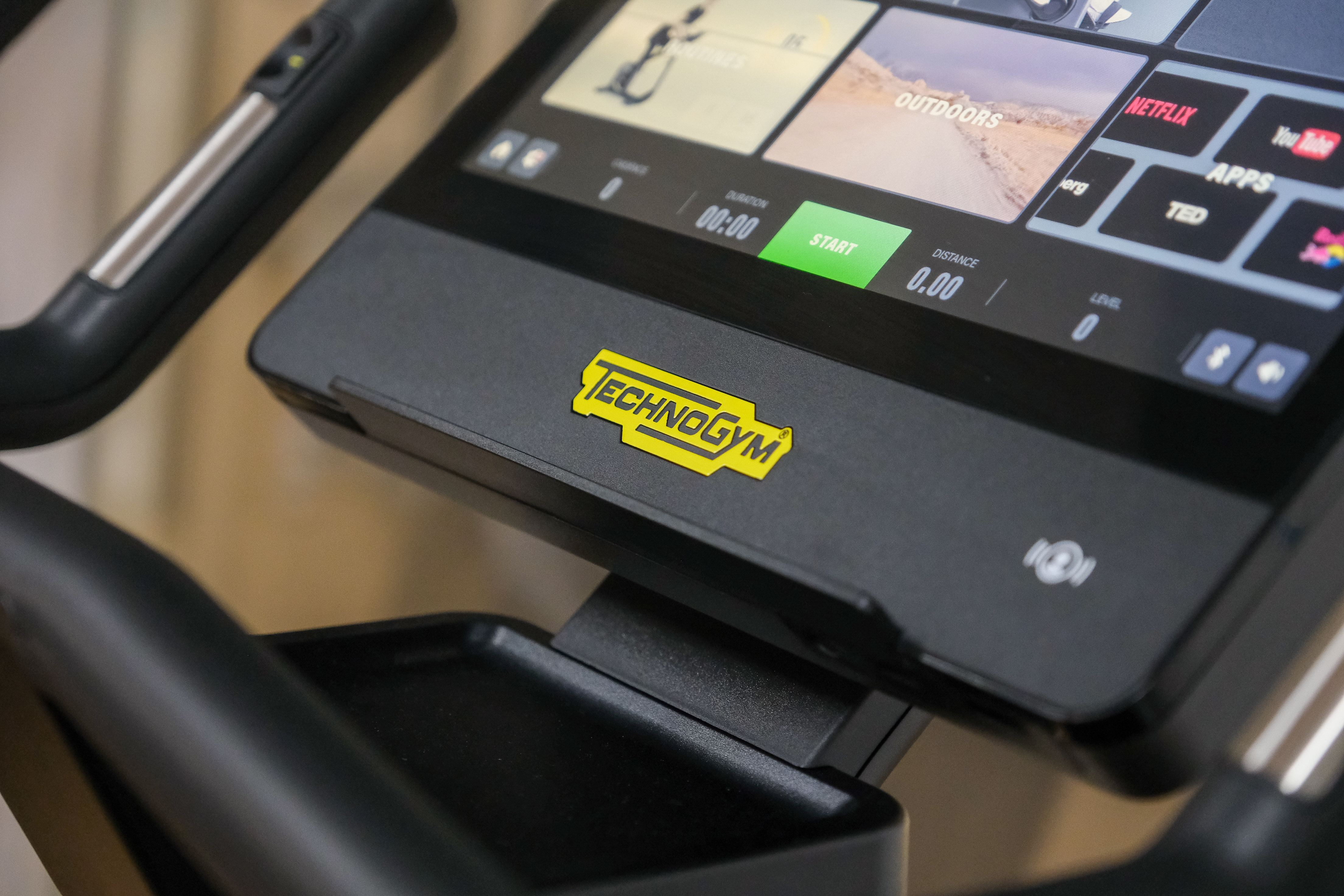 Close-up view of a Technogym exercise machine screen displaying workout options and apps, with the yellow Technogym logo visible on the console.