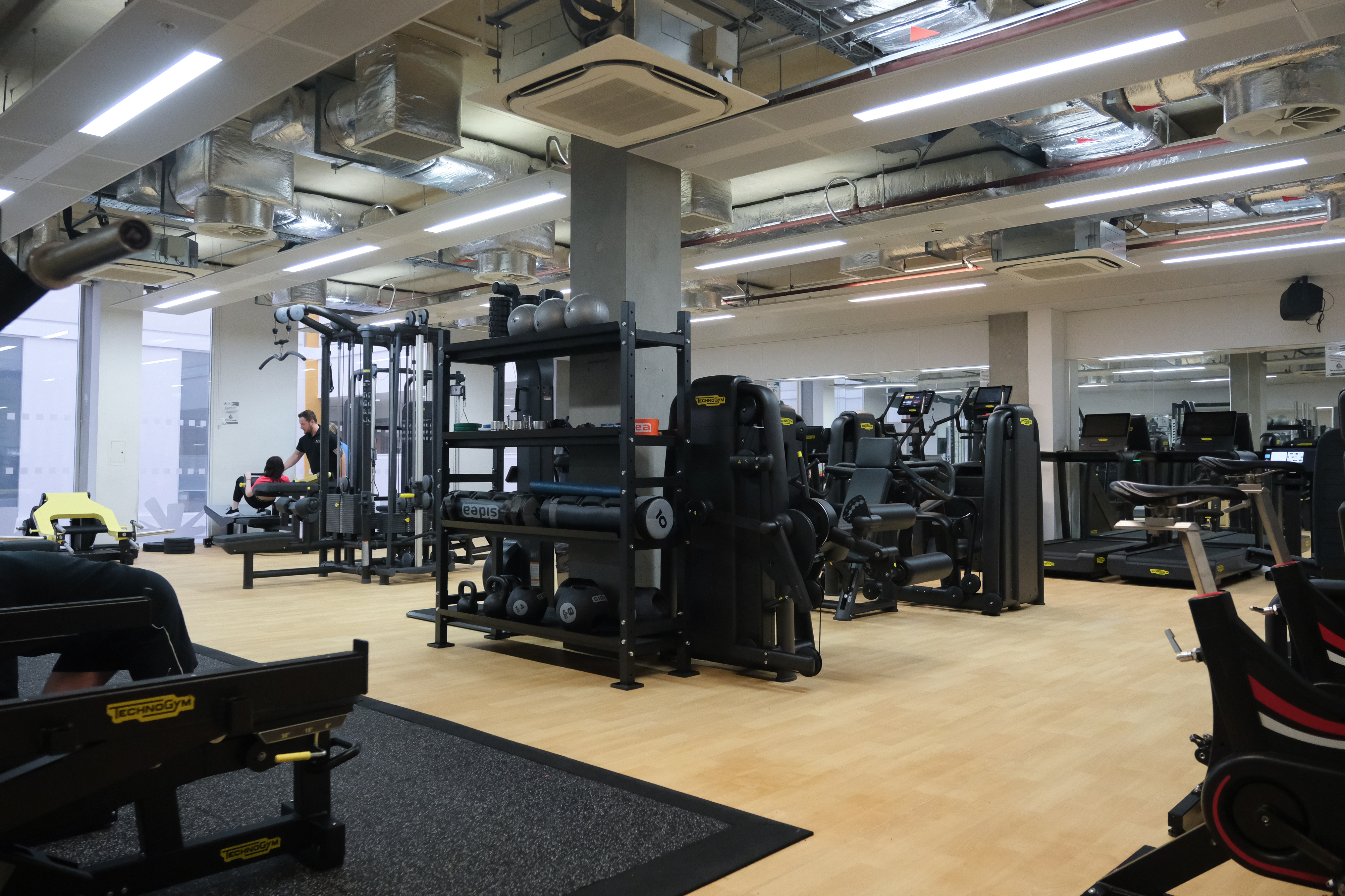 Modern gym interior with a variety of black fitness machines, weight racks, and benches arranged on a light wood floor. Exposed ductwork and lighting run across the ceiling, and mirrors line the back wall. A person is using equipment in the background, adding a sense of activity to the spacious workout area.
