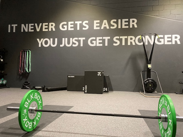 A gym wall with large white lettering that reads “IT NEVER GETS EASIER, YOU JUST GET STRONGER.” In front of the wall is a barbell loaded with green BLK BOX weight plates. Resistance bands hang on the left side of the wall, and a ski‑erg machine stands to the right. Soft plyo boxes are positioned along the back wall on grey rubber flooring.