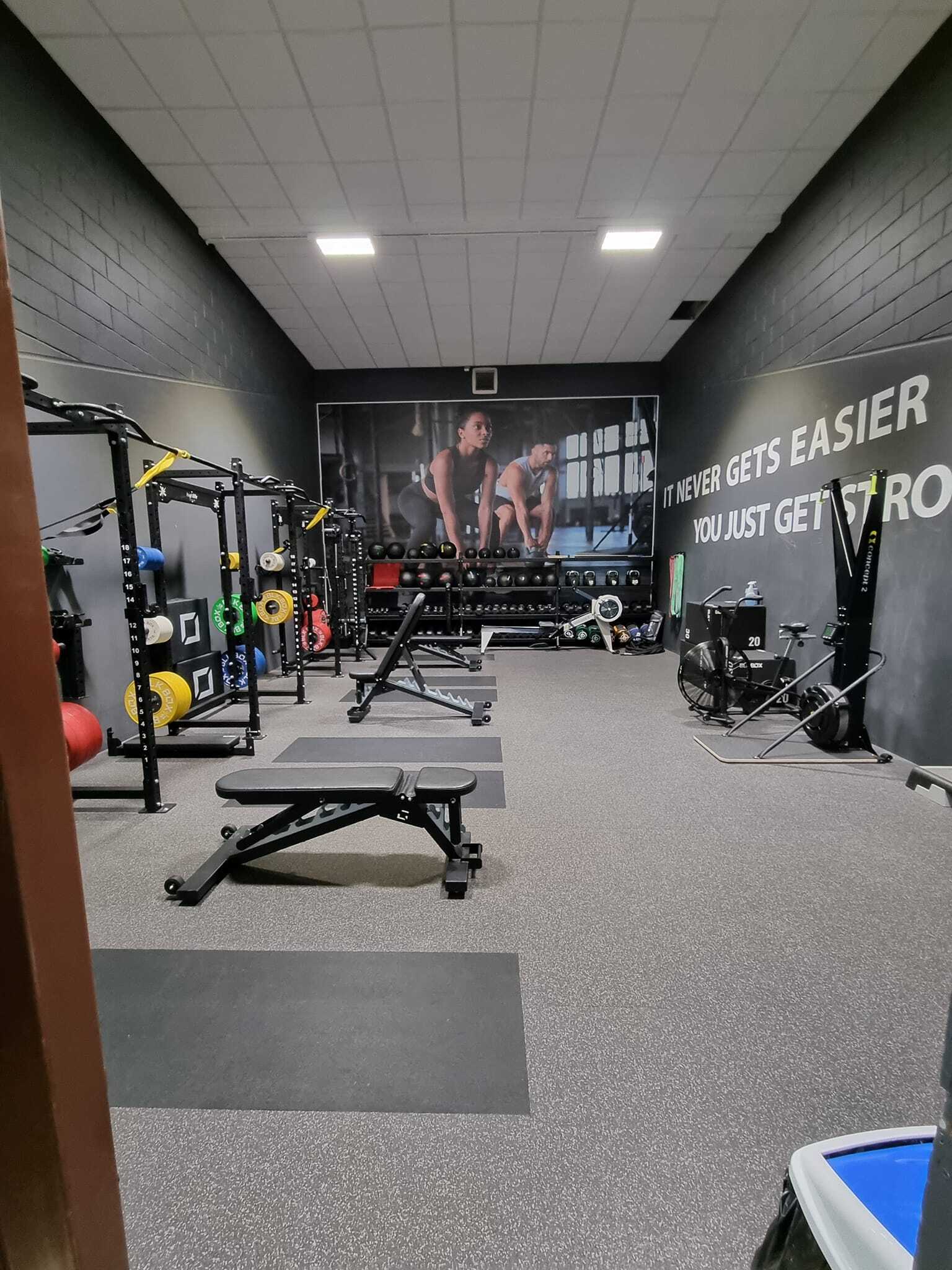 A compact strength‑training room with squat racks and colourful weight plates on the left, and adjustable benches positioned on rubber flooring. On the right wall, motivational text reads “IT NEVER GETS EASIER, YOU JUST GET STRONGER,” with exercise bikes and equipment beneath it. At the back of the room is a rack of dumbbells beneath a large wall mural showing people lifting weights.