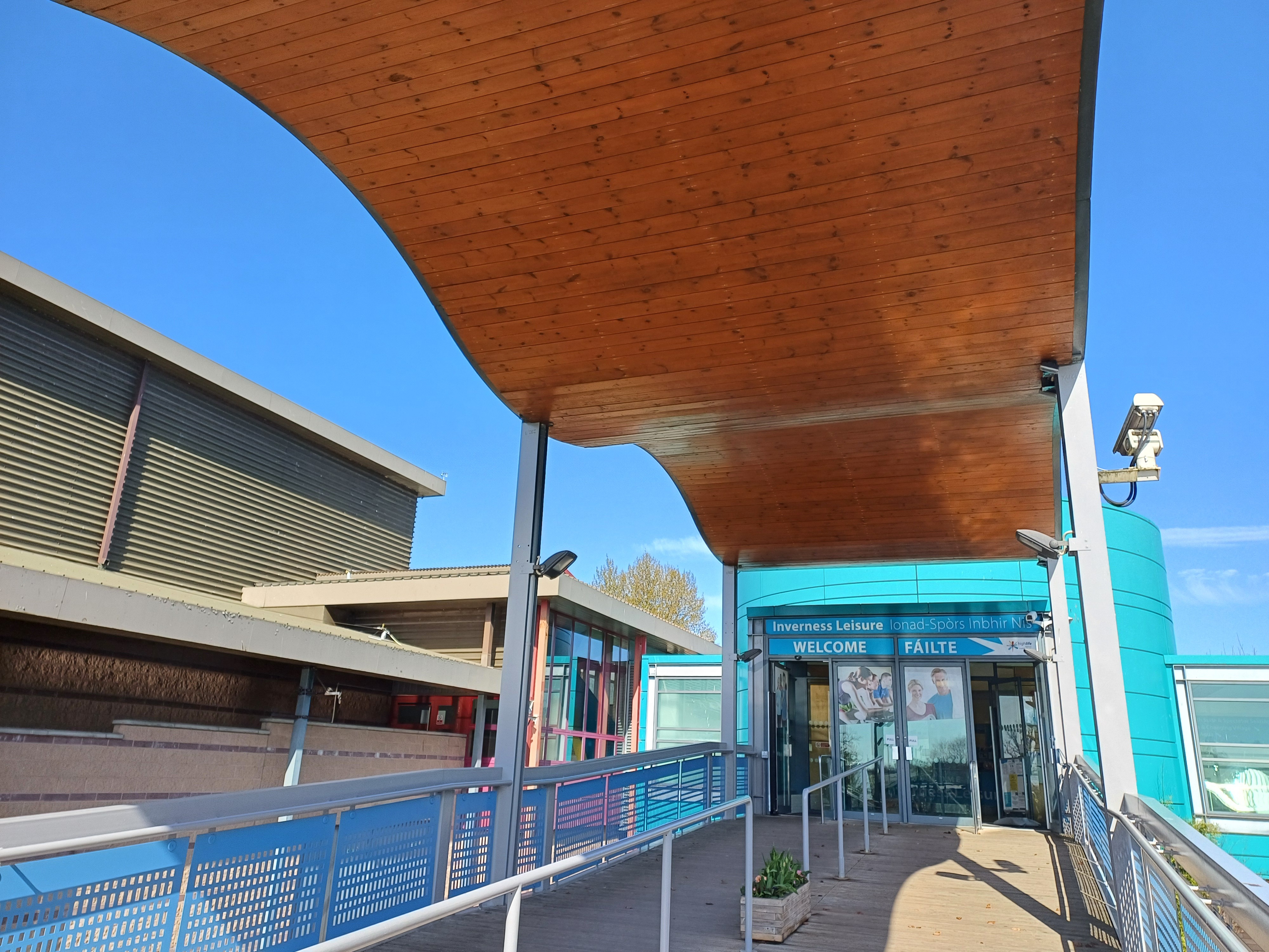 A walkway leads to the entrance of a leisure centre, with a large curved wooden canopy overhead and blue signage above the doors.