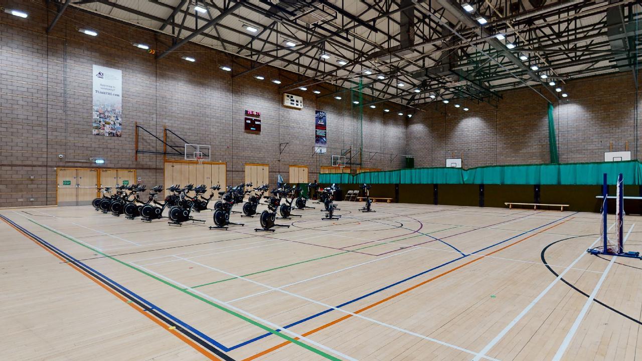 A large sports hall with rows of exercise bikes set up in the centre on a marked wooden court.