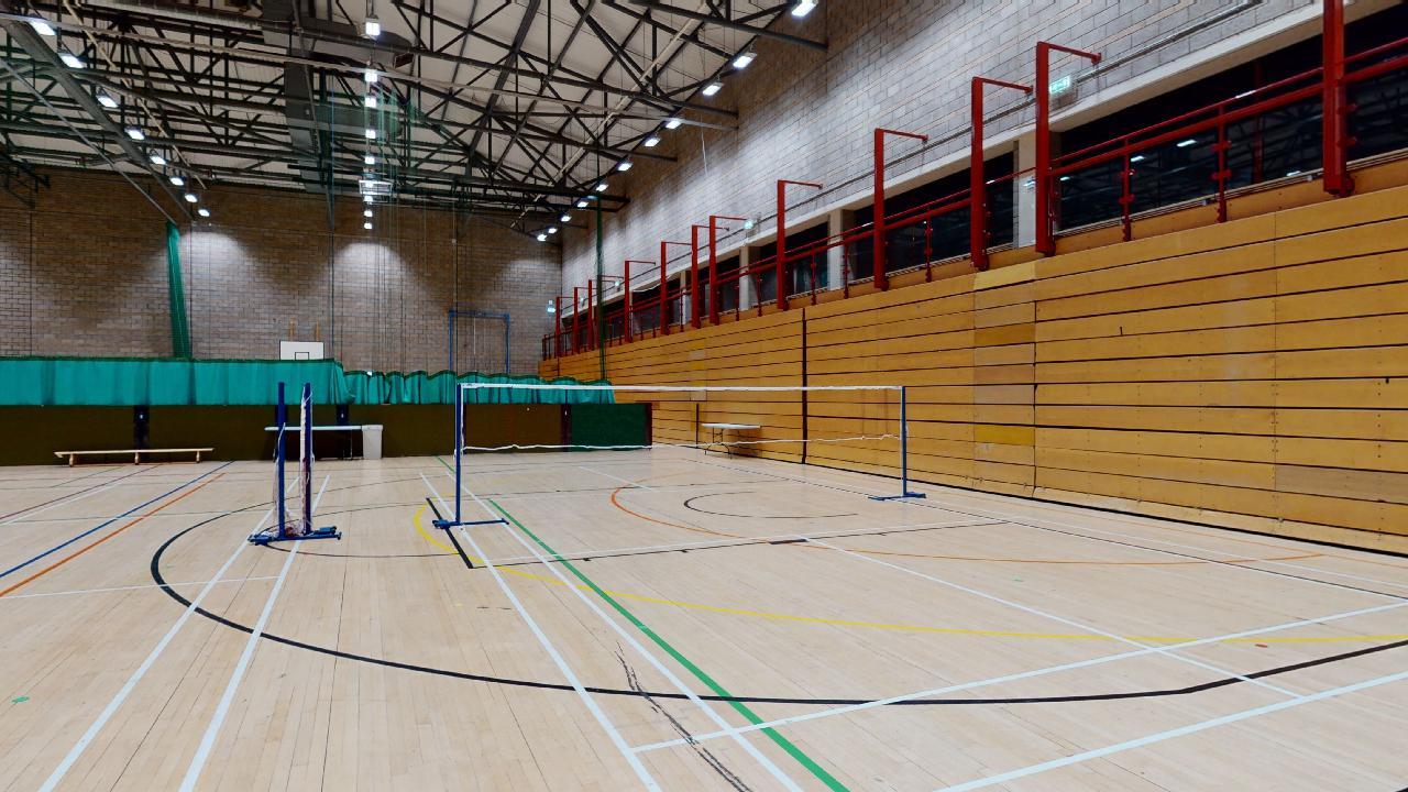 A sports hall with badminton nets set up on a marked wooden court, with tiered seating along one side.