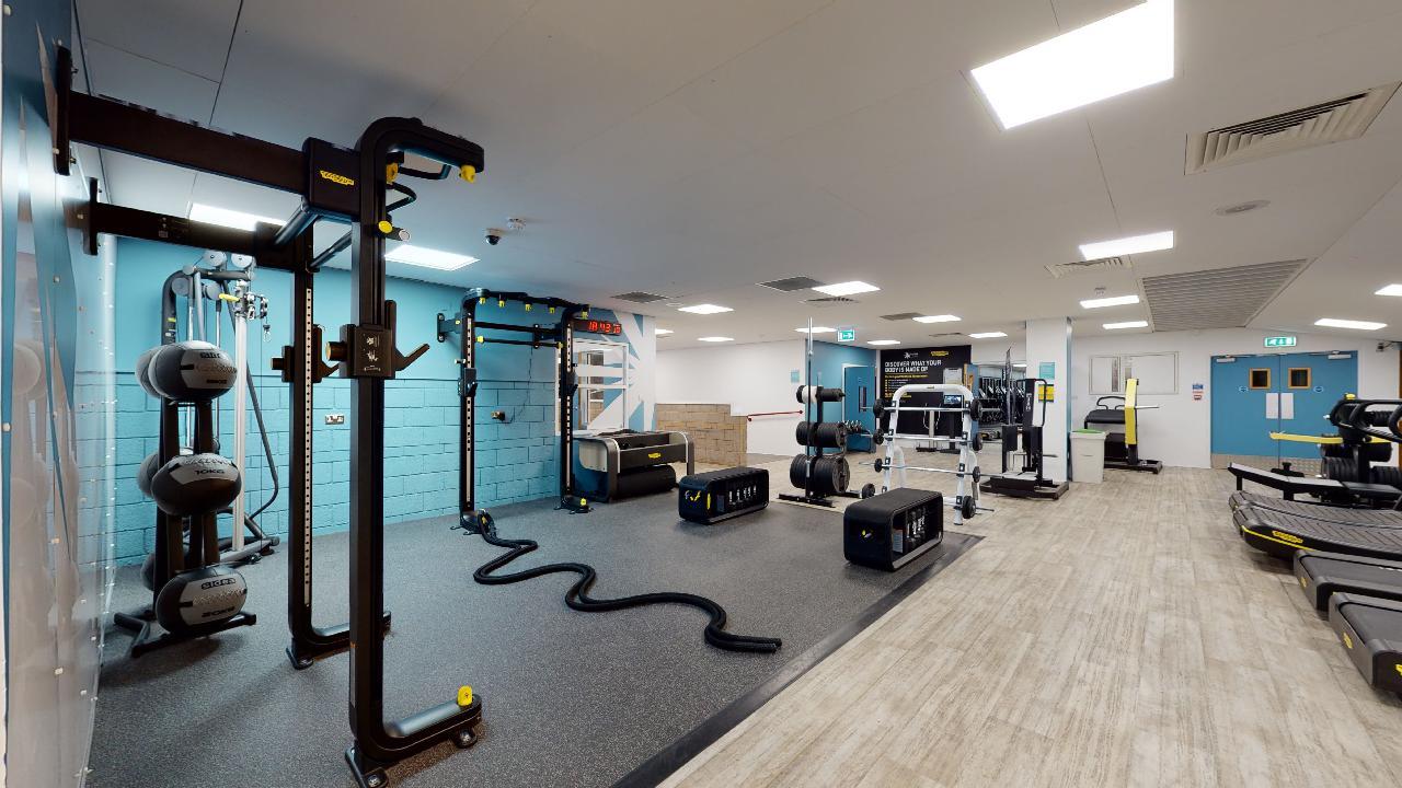 A spacious gym area featuring functional training equipment, including a multi‑station frame with attachments, battle ropes, weighted balls, and soft plyo boxes. Cardio machines such as treadmills are positioned on the right side, while weight benches and strength machines are arranged toward the back. The room has bright overhead lighting, blue and white walls, and a mix of rubber and wood‑effect flooring.
