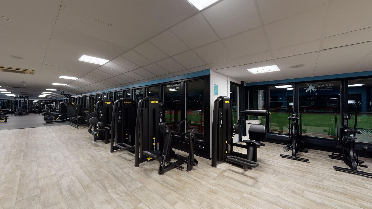 Strength machines and cardio equipment arranged beside large windows overlooking an outdoor area.