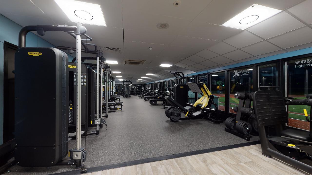 A gym area with resistance machines and other strength equipment arranged in rows beside large windows.