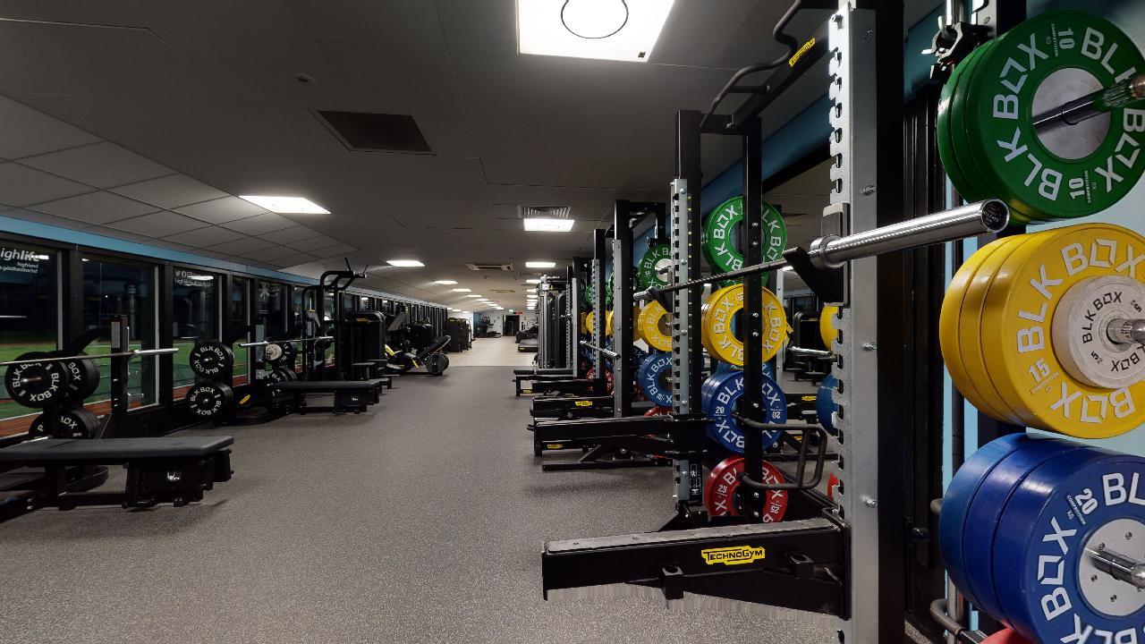 A spacious gym with weight racks, barbells and colourful weight plates, with benches and other strength equipment arranged in rows.