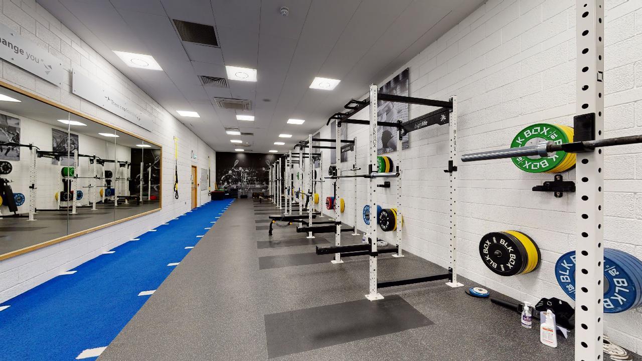 A long gym space lined with squat racks and colourful weight plates, with a blue turf track along one side.
