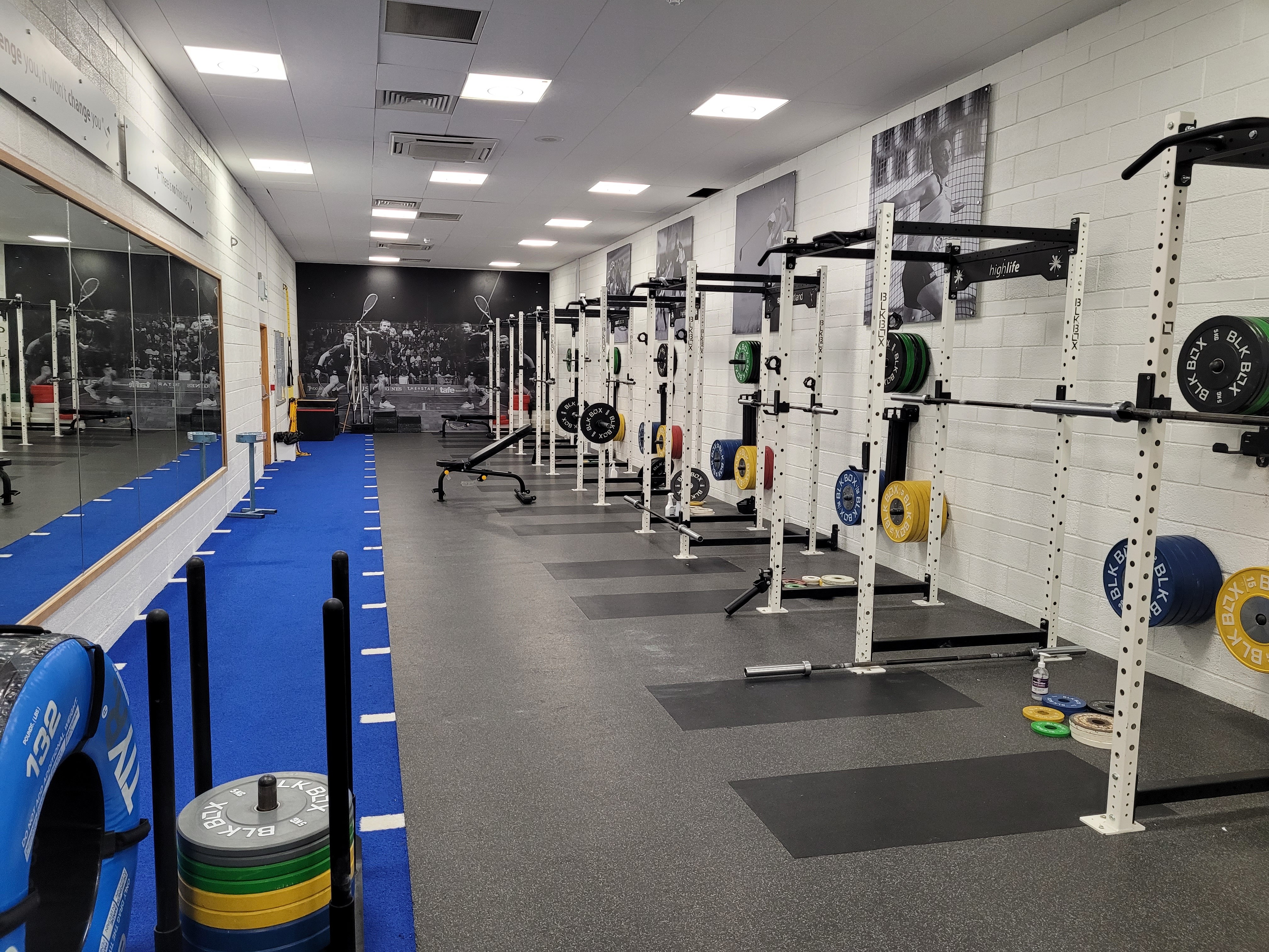 A long gym space lined with squat racks and colourful weight plates, with a blue turf track along one side.