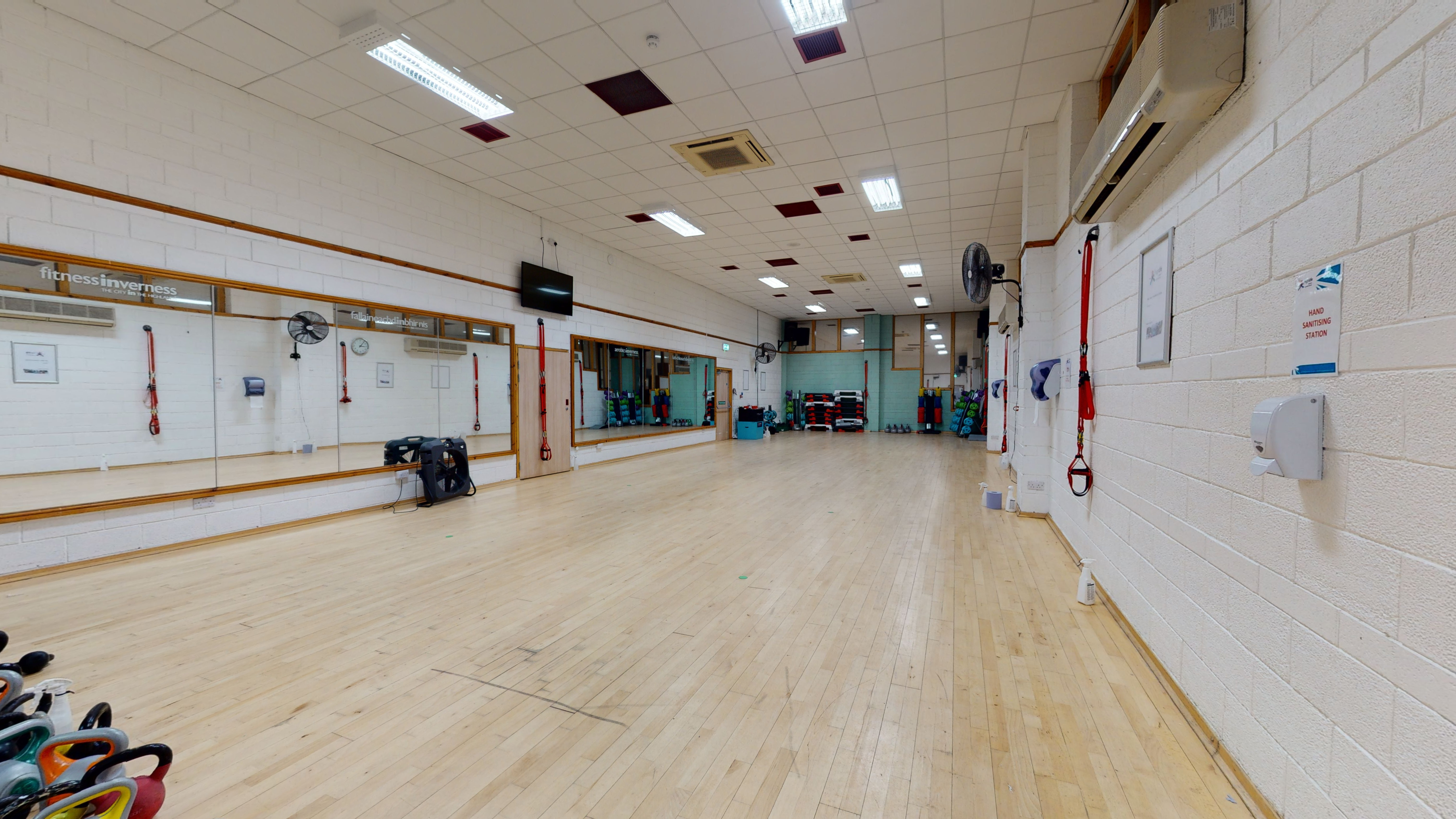 A spacious and well-lit studio featuring wooden flooring, with a set of kettlebells positioned in the foreground.