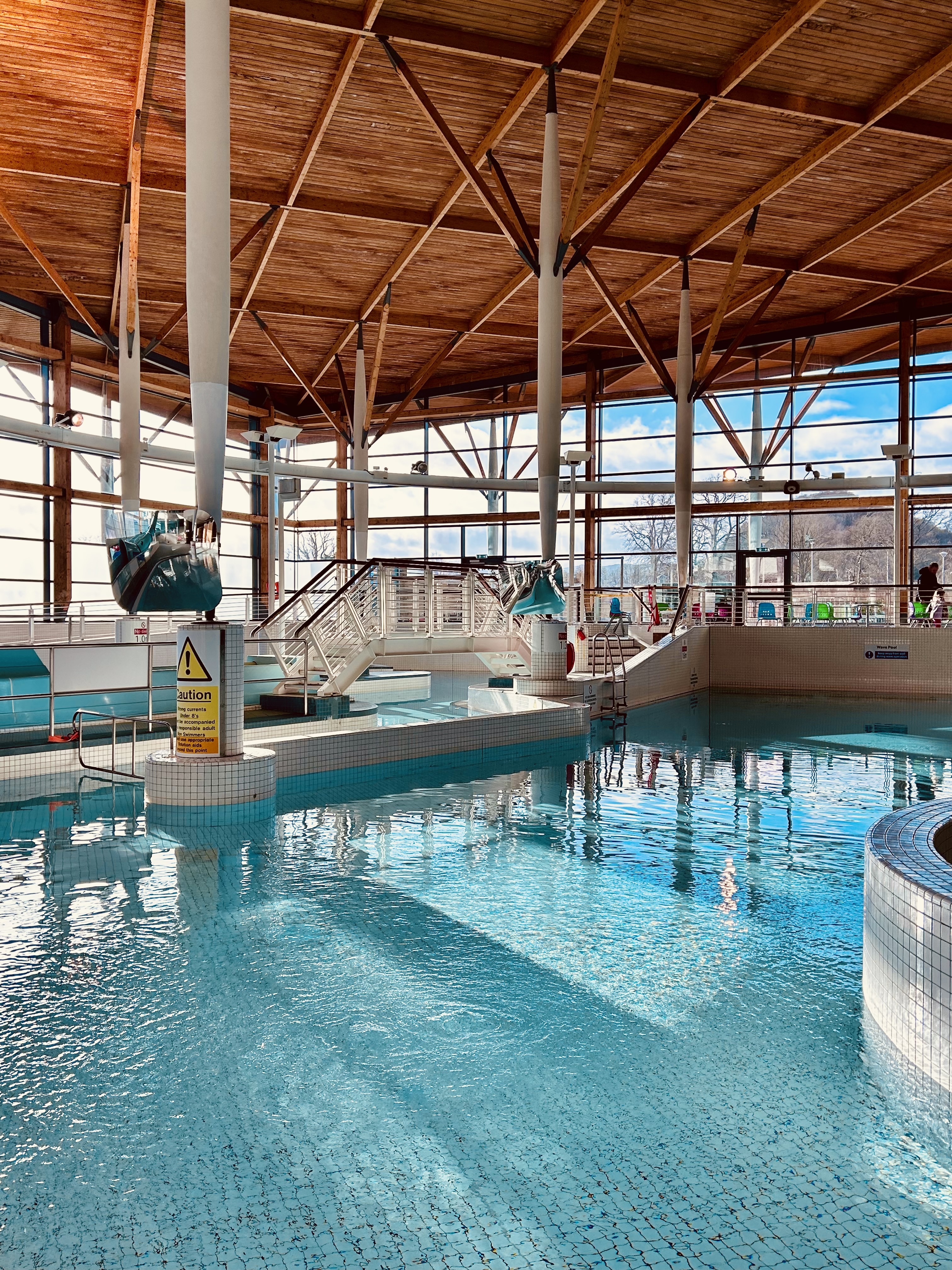 Bright indoor swimming pool with clear blue water, surrounded by white tiled edges. A raised walkway with railings crosses part of the pool, and water features shaped like dolphins are mounted on tall white pillars. Large floor‑to‑ceiling windows let in natural light, and the high wooden ceiling creates an open, airy feel.