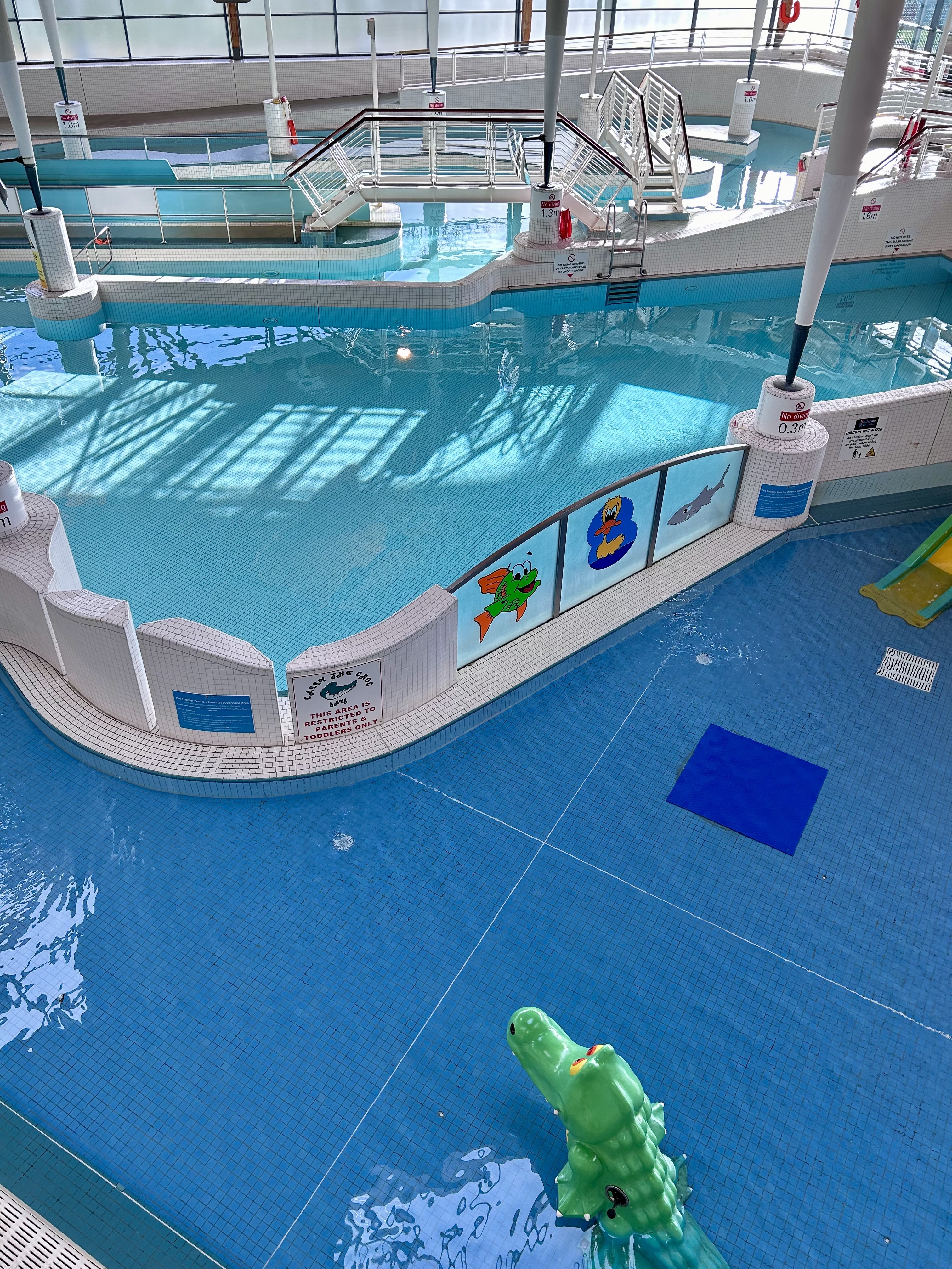 Indoor leisure pool with clear blue water, separated by low white tiled walls decorated with colourful aquatic-themed panels. A raised walkway with railings crosses part of the pool, leading to different water areas. Large floor‑to‑ceiling windows surround the space, letting in natural light, and a high wooden ceiling supported by white pillars creates an open, bright atmosphere.
