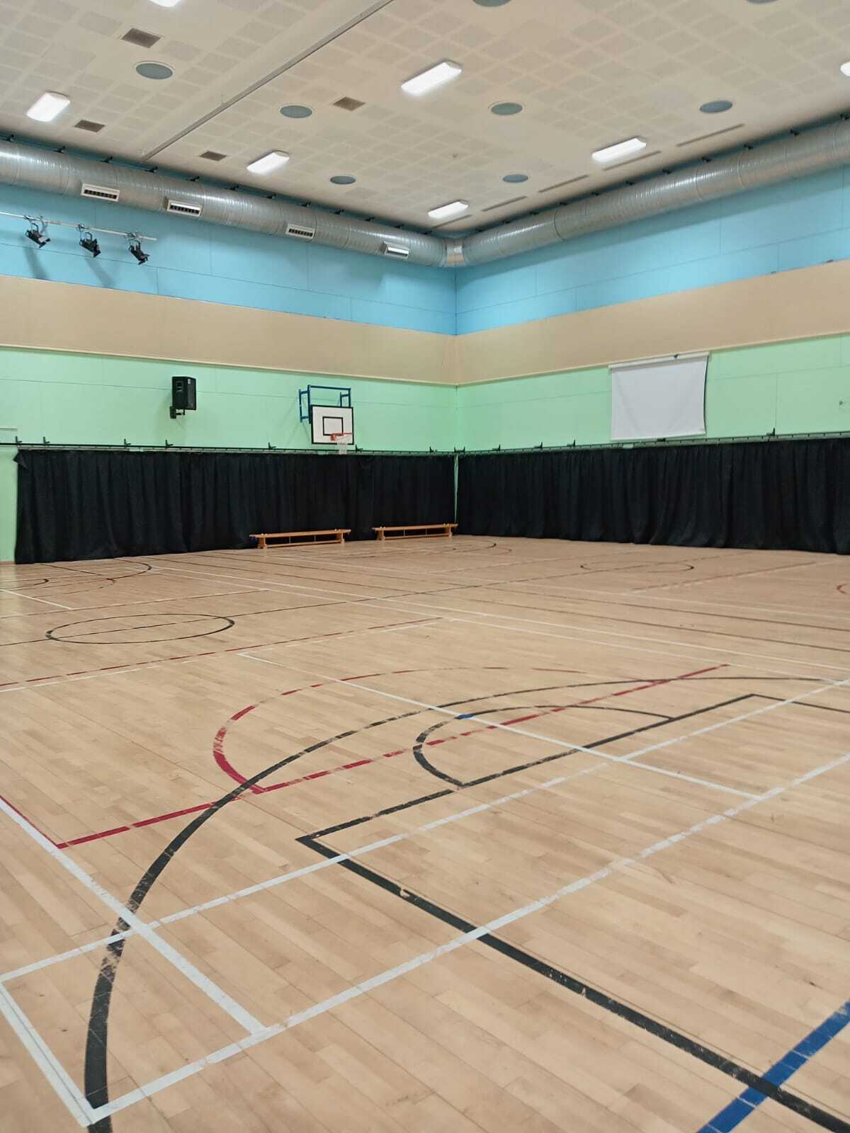 Spacious indoor sports hall with a polished wooden floor marked for multiple games, including basketball. Two basketball hoops are mounted on the mint‑green and blue walls, and black curtains line one side of the hall. Bright ceiling lights and exposed ventilation ducts give the space an open, modern feel.
