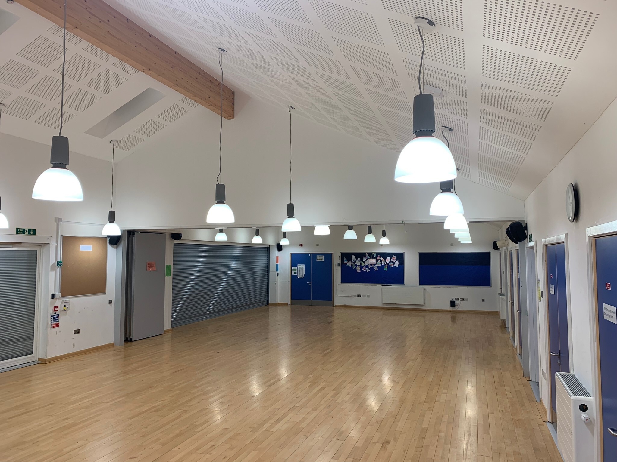 Bright community room with a polished wooden floor, white walls and a high, angled ceiling fitted with hanging dome lights. Several blue doors line the right wall, and metal roller shutters cover service areas on the left. A noticeboard and display wall are visible at the far end of the spacious, multi‑use room.