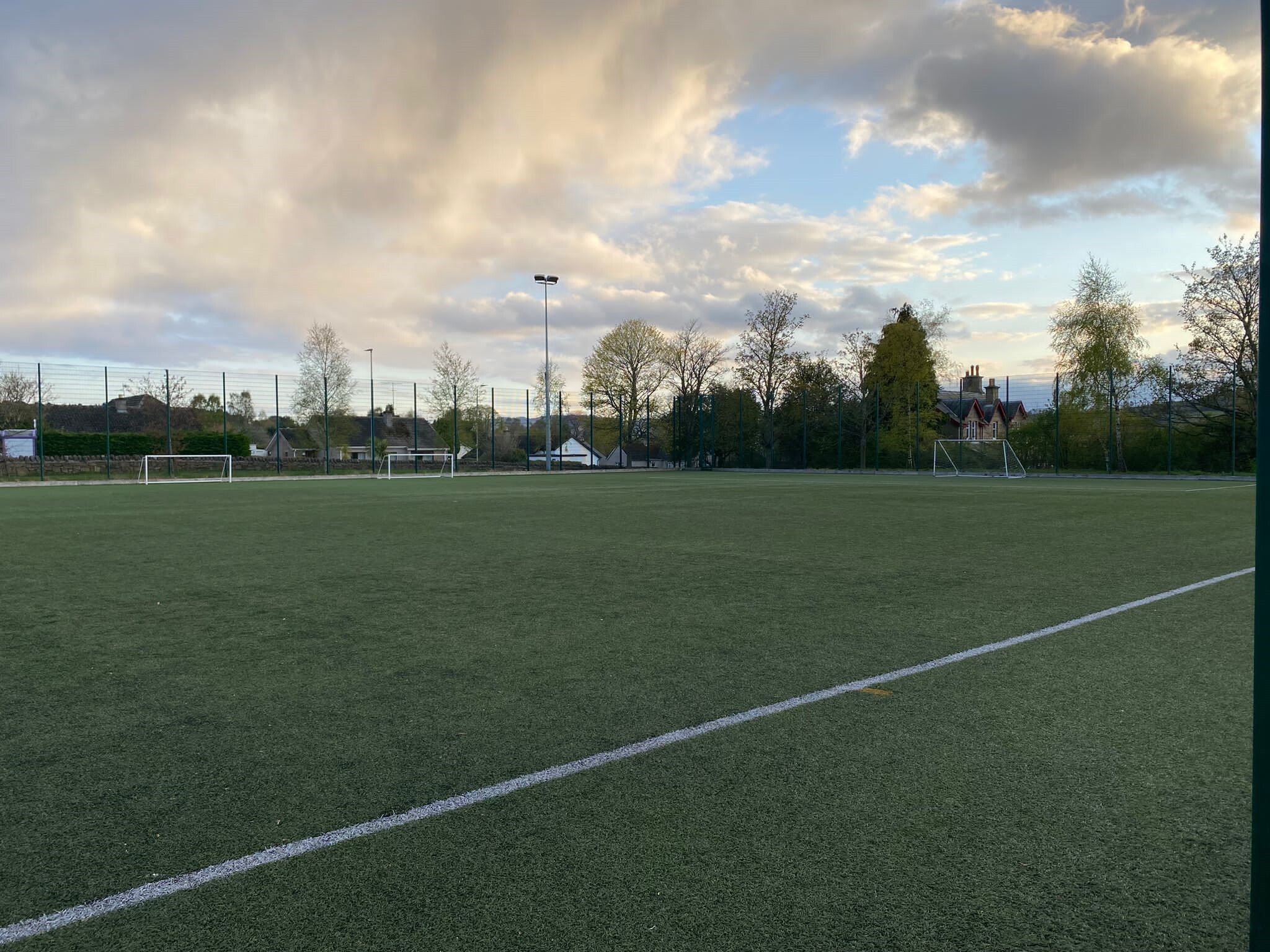 Outdoor artificial turf football pitch with white line markings and several small goalposts in the distance. The pitch is surrounded by tall fencing, with trees and houses beyond it. The sky is partly cloudy with soft evening light.