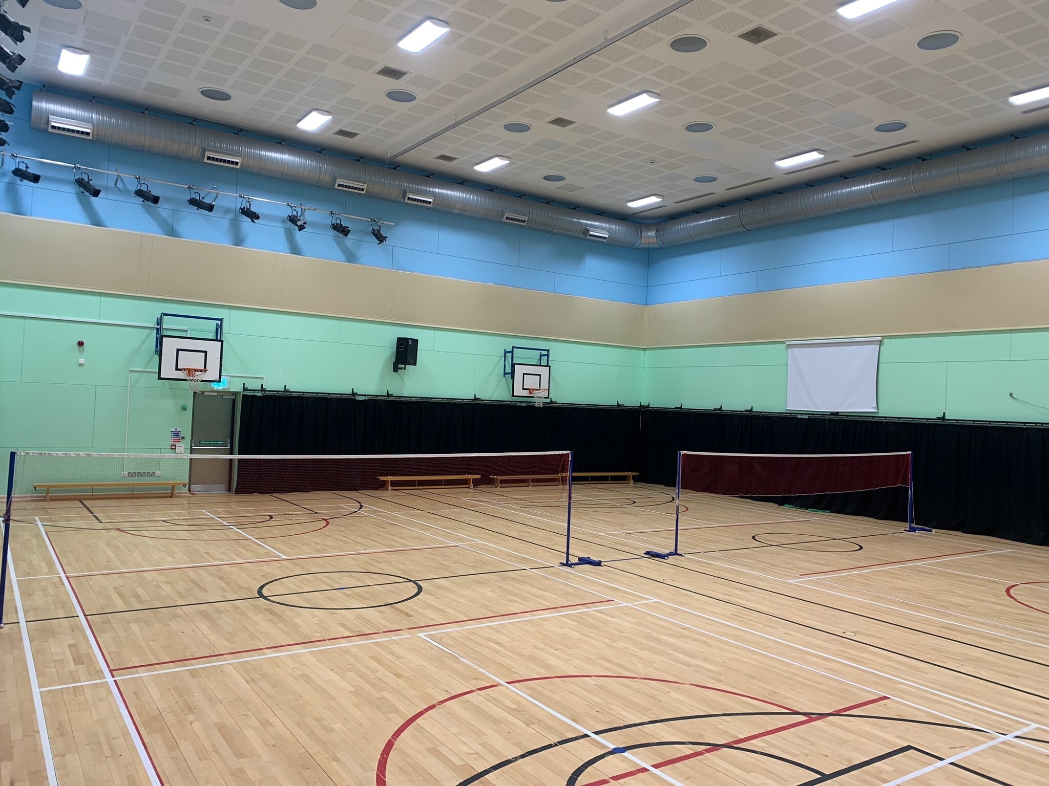 Indoor sports hall with a polished wooden floor marked for multiple games, including basketball and badminton. Two badminton courts are set up with nets, and basketball hoops are mounted on the walls. The walls are painted in blue and green panels, and bright ceiling lights illuminate the large, spacious hall.