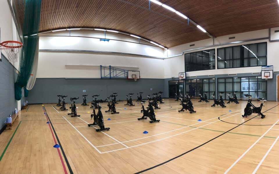 A spacious games hall featuring floor markings for basketball and badminton courts, equipped with basketball hoops mounted on the walls, and arranged to accommodate an indoor cycling class.