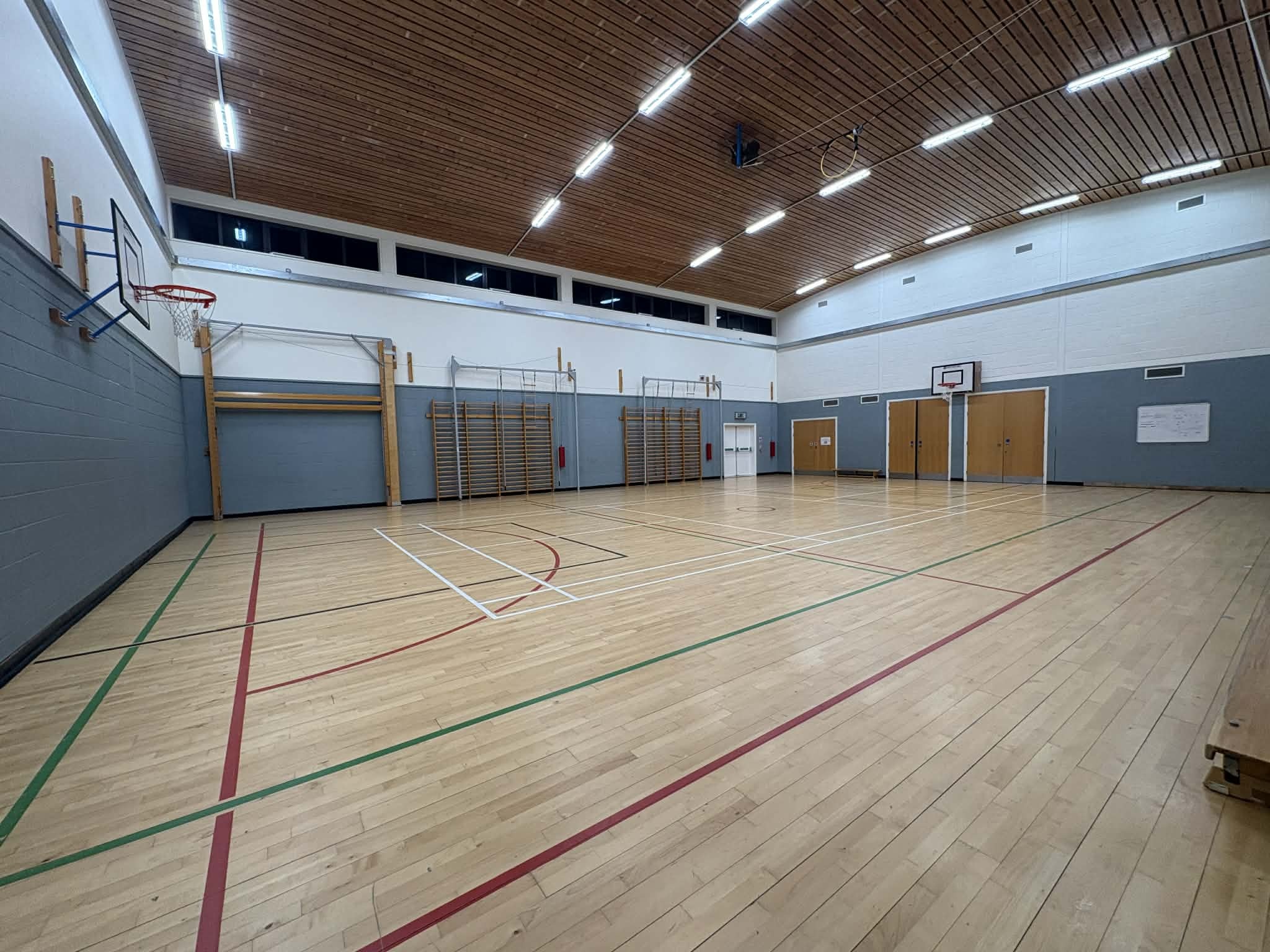 A well-lit hall featuring floor markings for both basketball and badminton, along with a basketball hoop mounted on the wall.