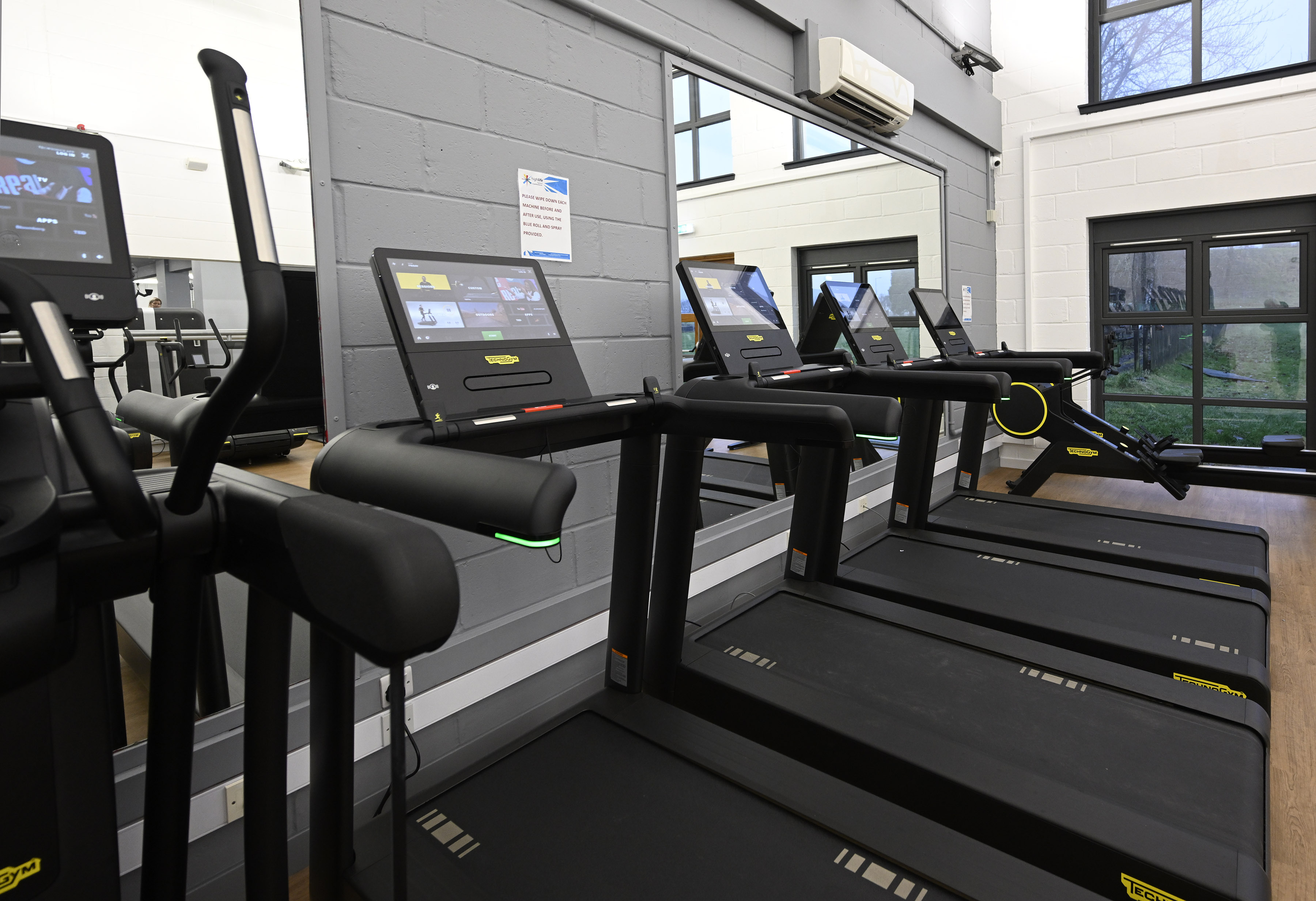 Row of Technogym treadmills in a bright gym, positioned in front of a large mirrored wall. Each treadmill has a touchscreen display, and the space features grey and white walls, large windows, and wooden flooring.