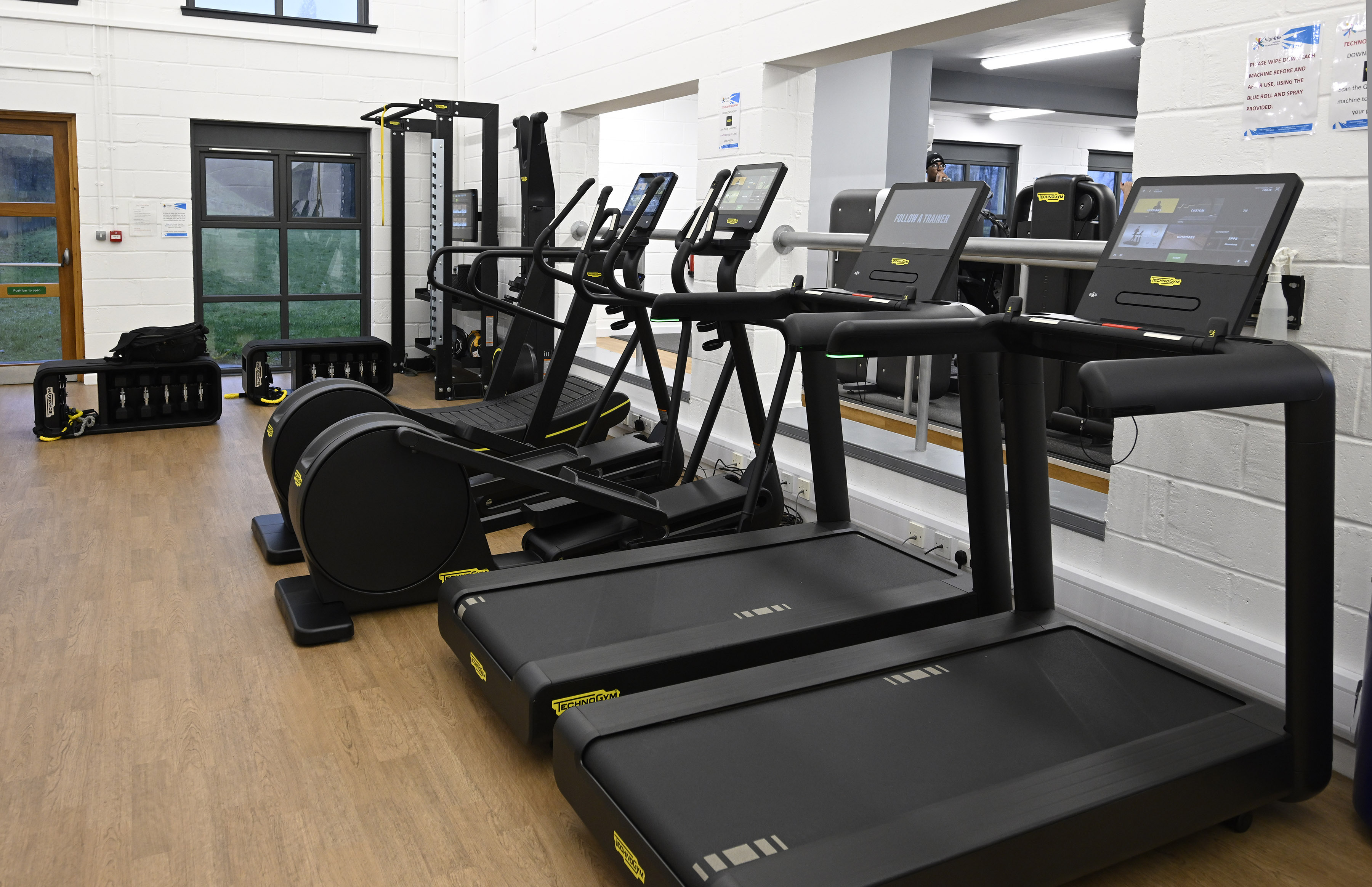 Modern gym space with a row of Technogym treadmills and other cardio machines facing mirrored walls. The room has light wooden flooring, white brick walls, and large windows letting in natural light. Additional strength‑training equipment is arranged toward the back of the room.