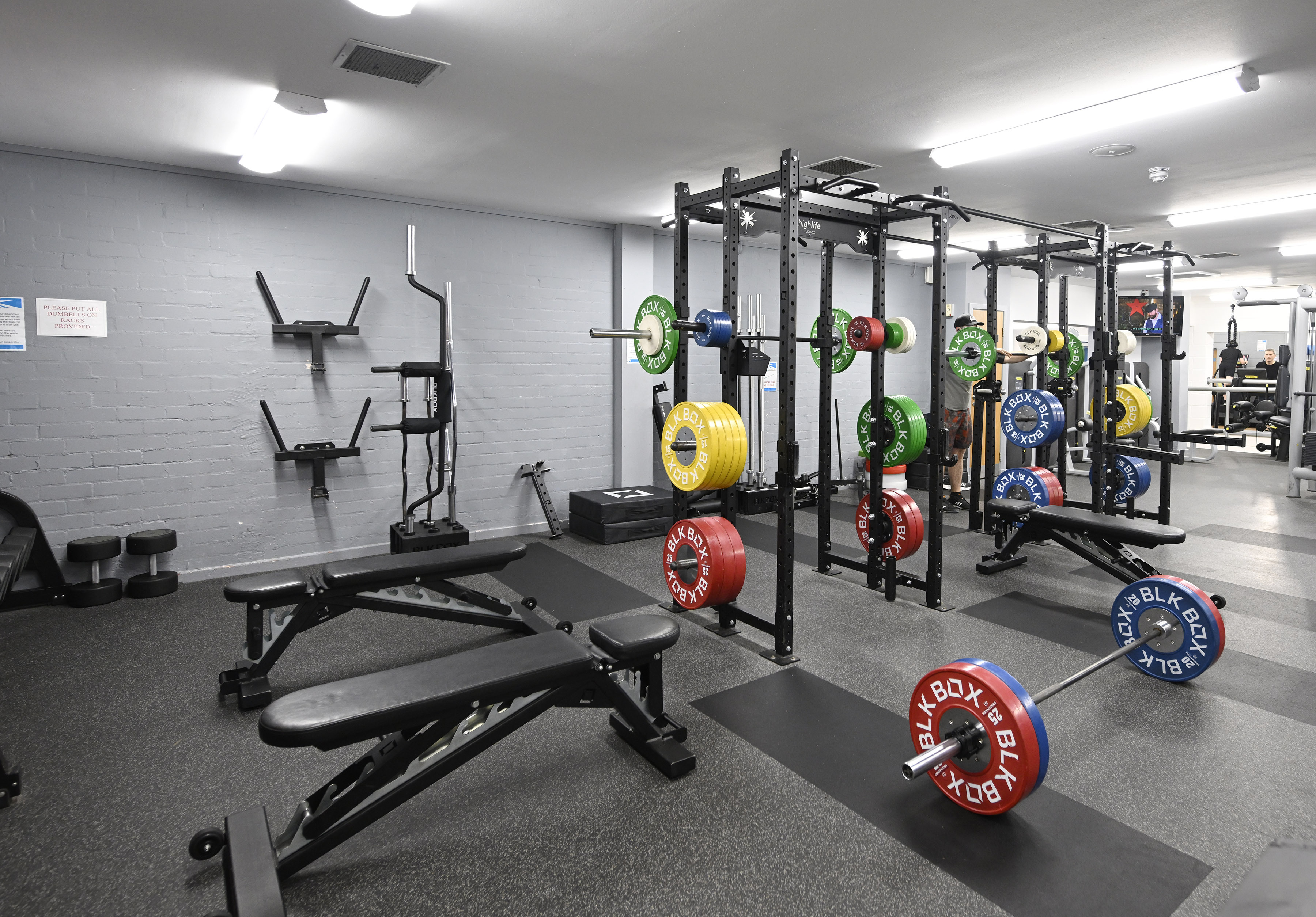 Strength‑training room with black weight benches, a power rack fitted with colourful weight plates, and barbells on the floor. Additional equipment, including wall‑mounted storage and squat stands, is set against a grey brick wall. The space is well‑lit with bright ceiling lights and has a rubber gym floor.