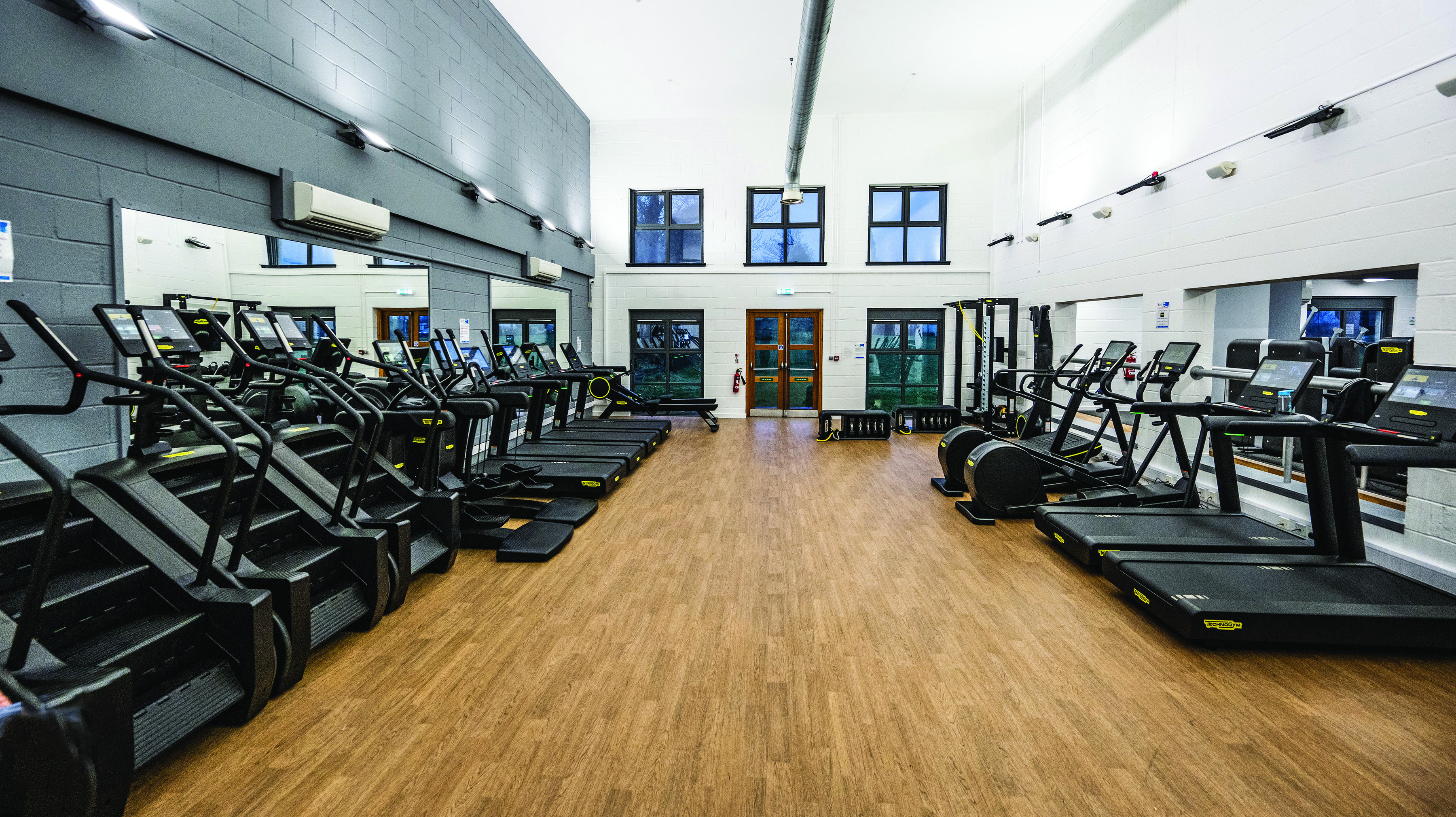 Spacious, modern gym with rows of Technogym cardio equipment, including treadmills and stair climbers, arranged along both sides of the room. The gym has light wooden flooring, large mirrors on one wall, high ceilings with exposed ducting, and tall windows and double doors at the far end letting in natural light