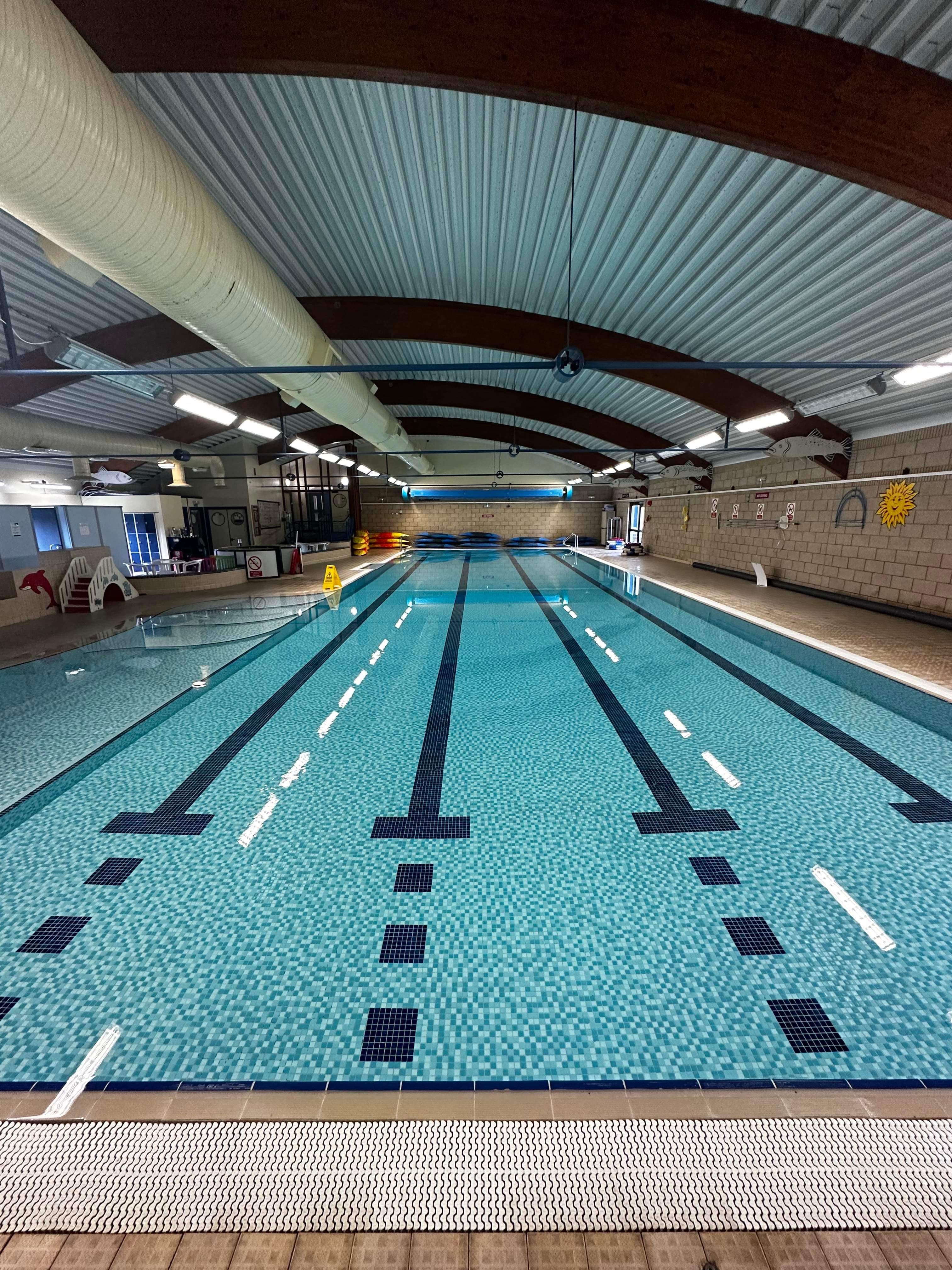 Indoor swimming pool with six lanes, clear blue water, lane dividers, and exposed ceiling beams with ventilation ducts.