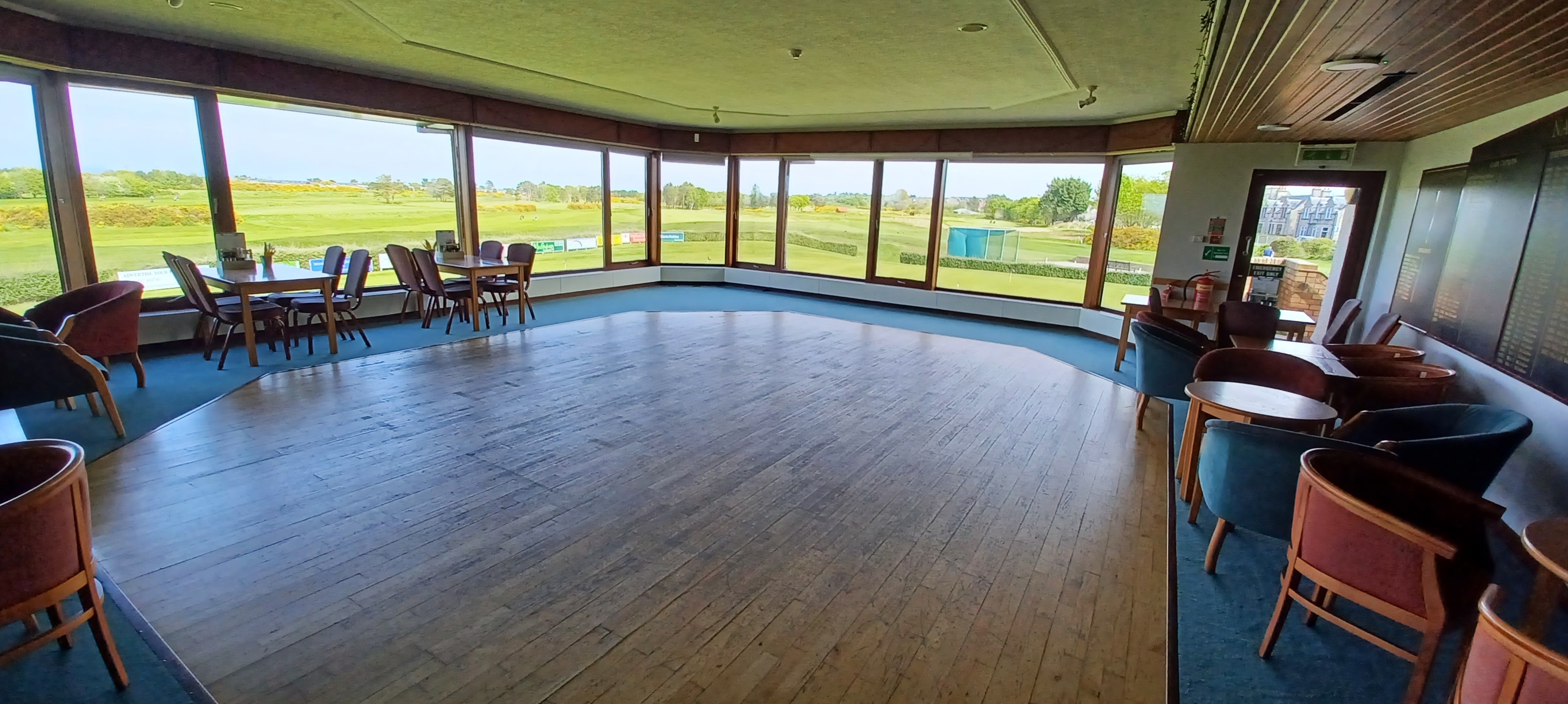 A spacious room featuring hardwood flooring and tables and chairs arranged along the periphery. Floor-to-ceiling windows offer stunning views of the golf course.