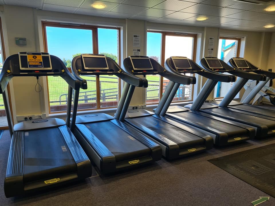Row of Technogym treadmills in a bright gym, positioned in front of large windows overlooking an outdoor sports field. The treadmills have digital screens, and the room is lit with ceiling spotlights.