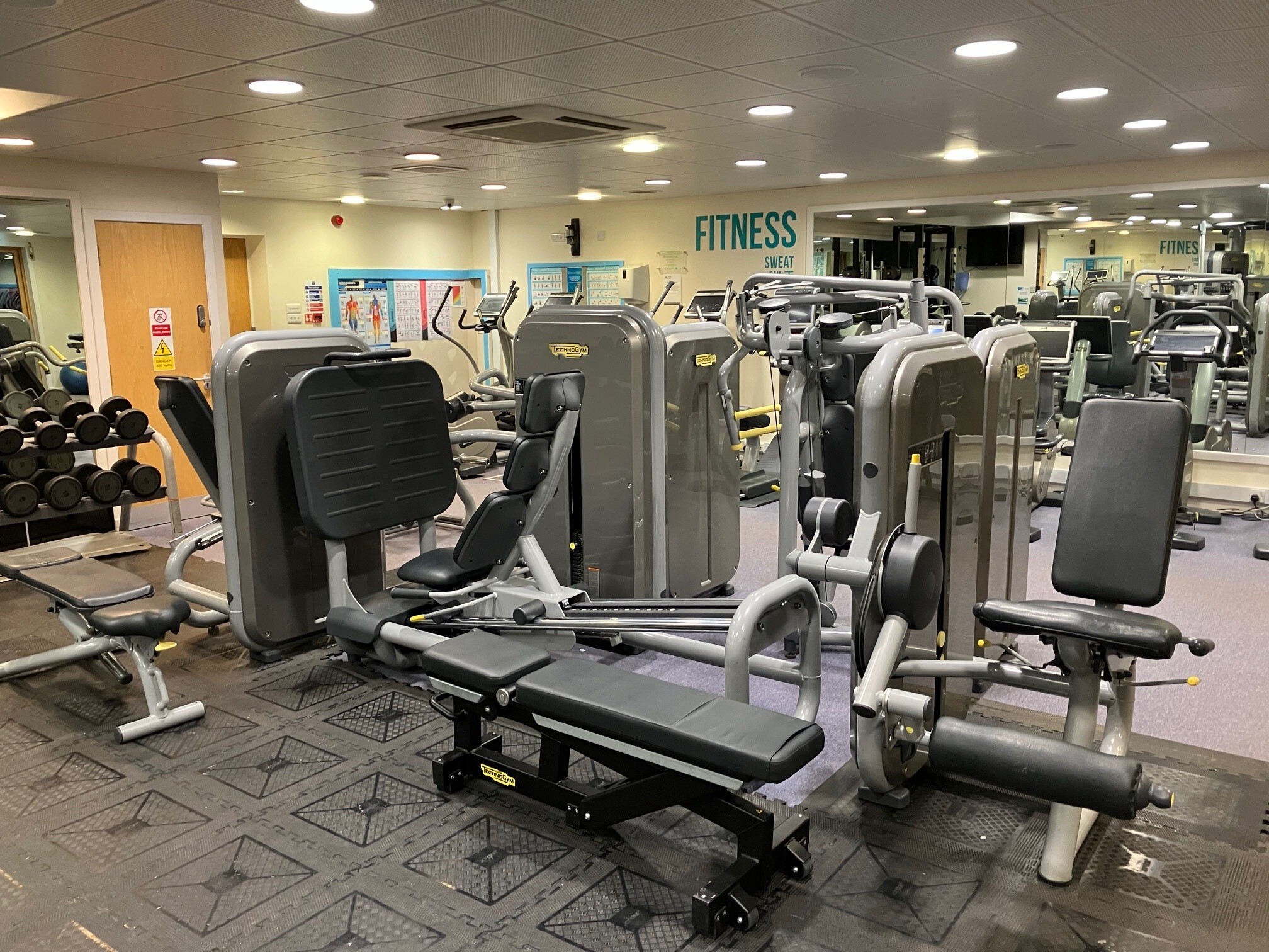 Gym area with several Technogym resistance machines, including a leg press, leg extension, and adjustable bench. A rack of dumbbells sits to the left beside mirrored walls, and bright ceiling lights illuminate the spacious room. Motivational text reading ‘Fitness’ is visible on the far wall.