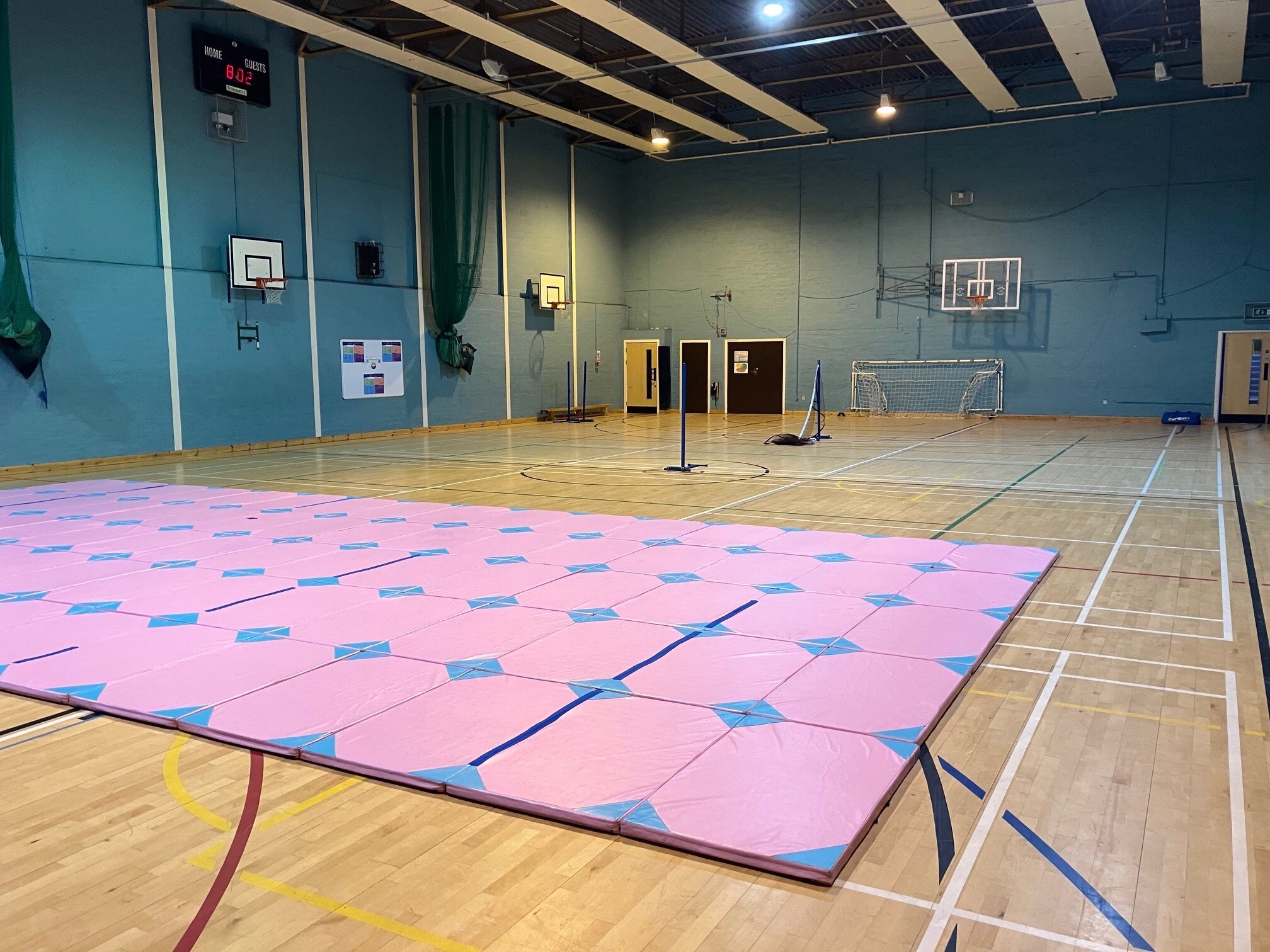 Large games hall featuring basketball and badminton court floor markings, basketball hoops on the walls and a goal against the end wall. There is a large colourful fitness matt on the floor within the first half of the hall, with badminton nets set up within the second half of the hall.